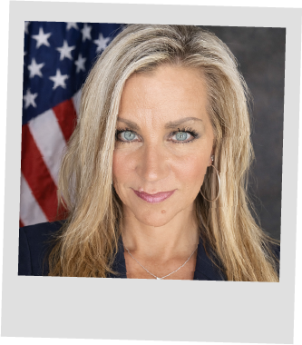 Portrait of a blonde woman with blue eyes wearing hoop earrings in front of an American flag.
