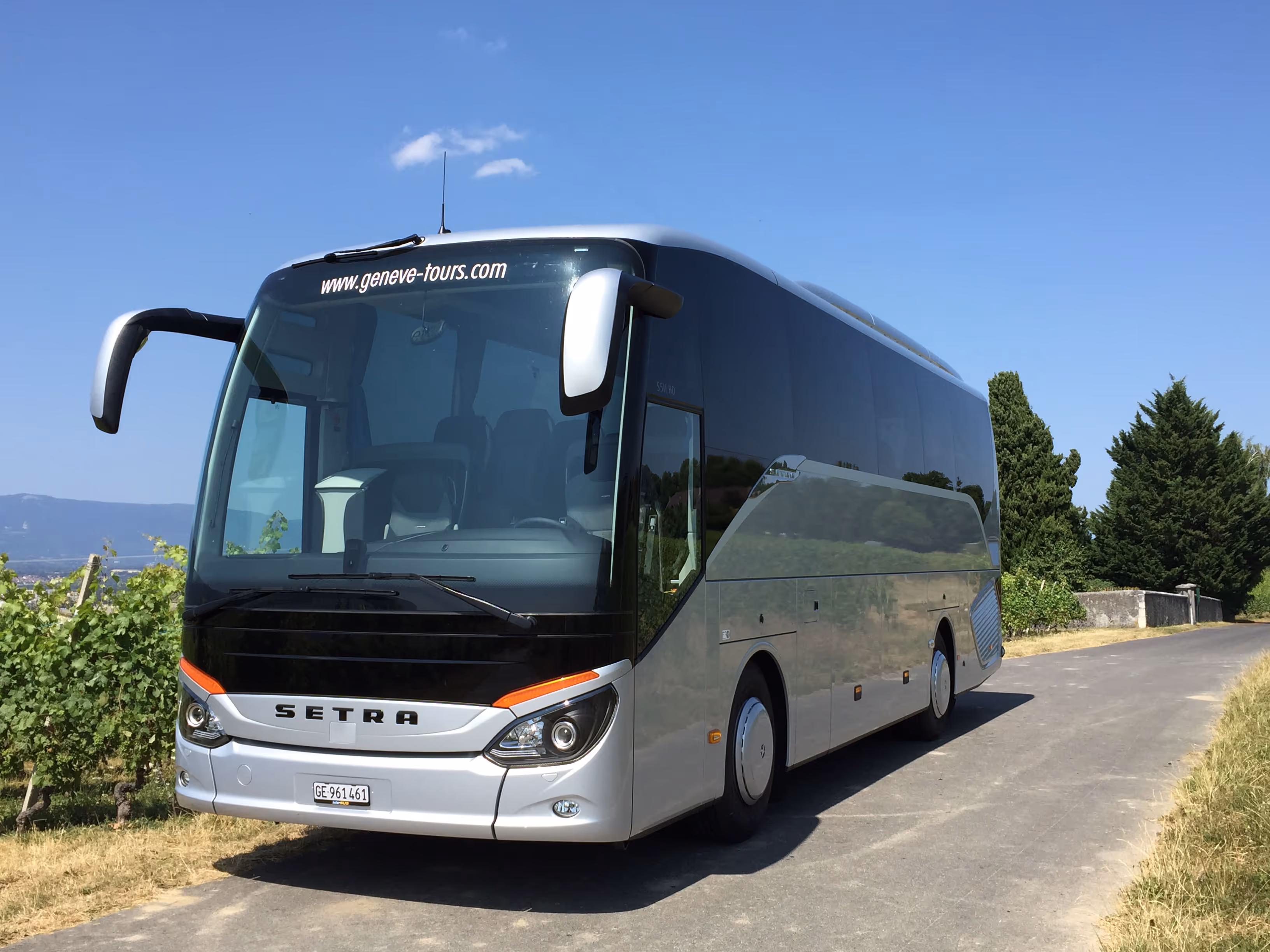 Vue extérieure de trois-quarts d'un autocar de tourisme Setra gris métallisé, stationné sur une route de campagne bordée de vignes sous un ciel bleu.