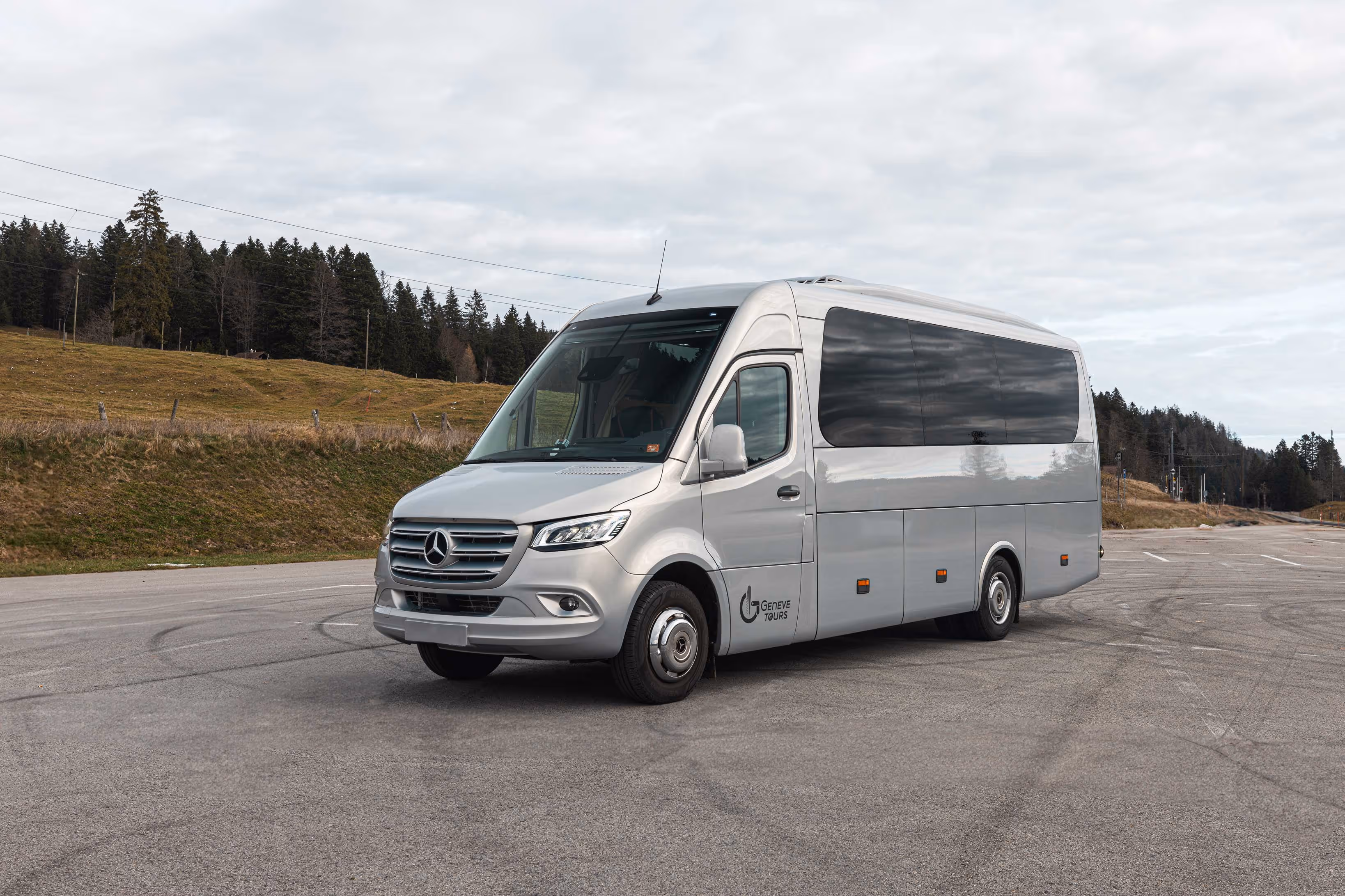 Minibus Mercedes-Benz argenté de Geneve Tours stationné sur un parking, avec des vitres teintées noires et une forêt en arrière-plan