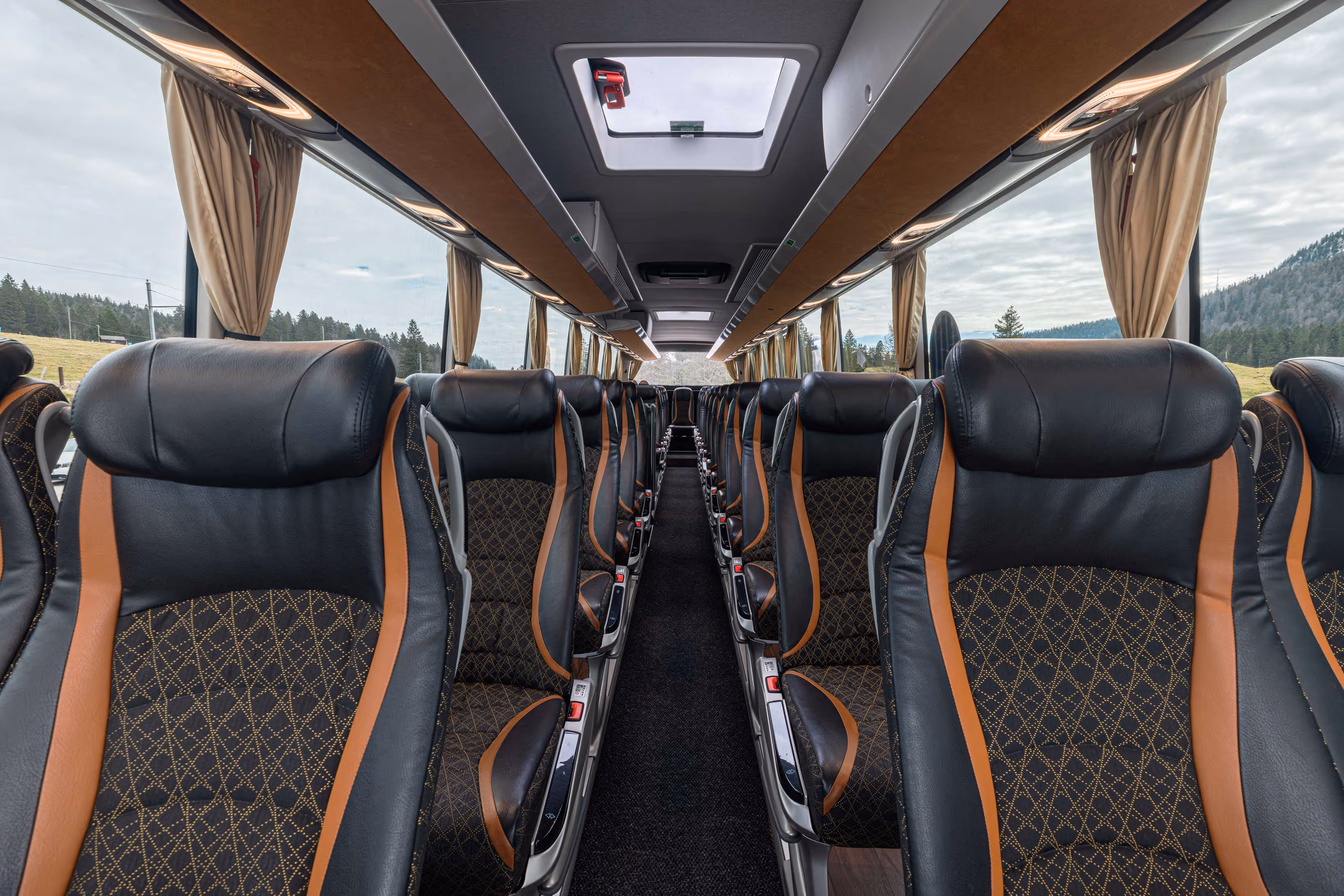 Perspective intérieure de l'allée centrale d'un autocar, dévoilant des sièges en cuir noir avec des finitions marron et des motifs losanges, sous un plafond équipé de trappes de ventilation lumineuses.