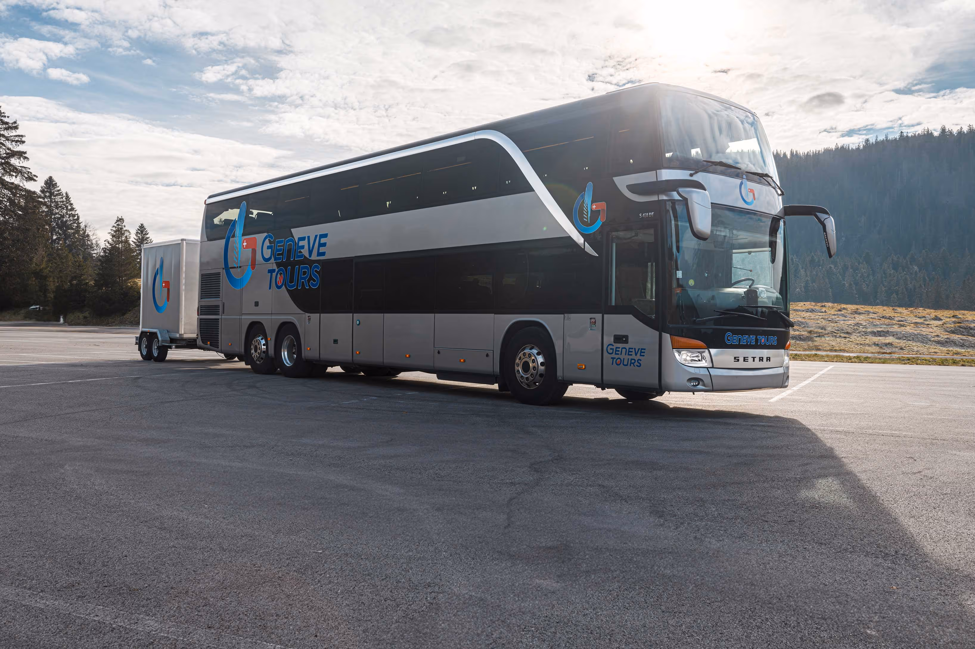 Autocar de tourisme Setra à deux étages gris métallisé, floqué du logo Geneve Tours, tractant une remorque assortie sur un grand parking dégagé.