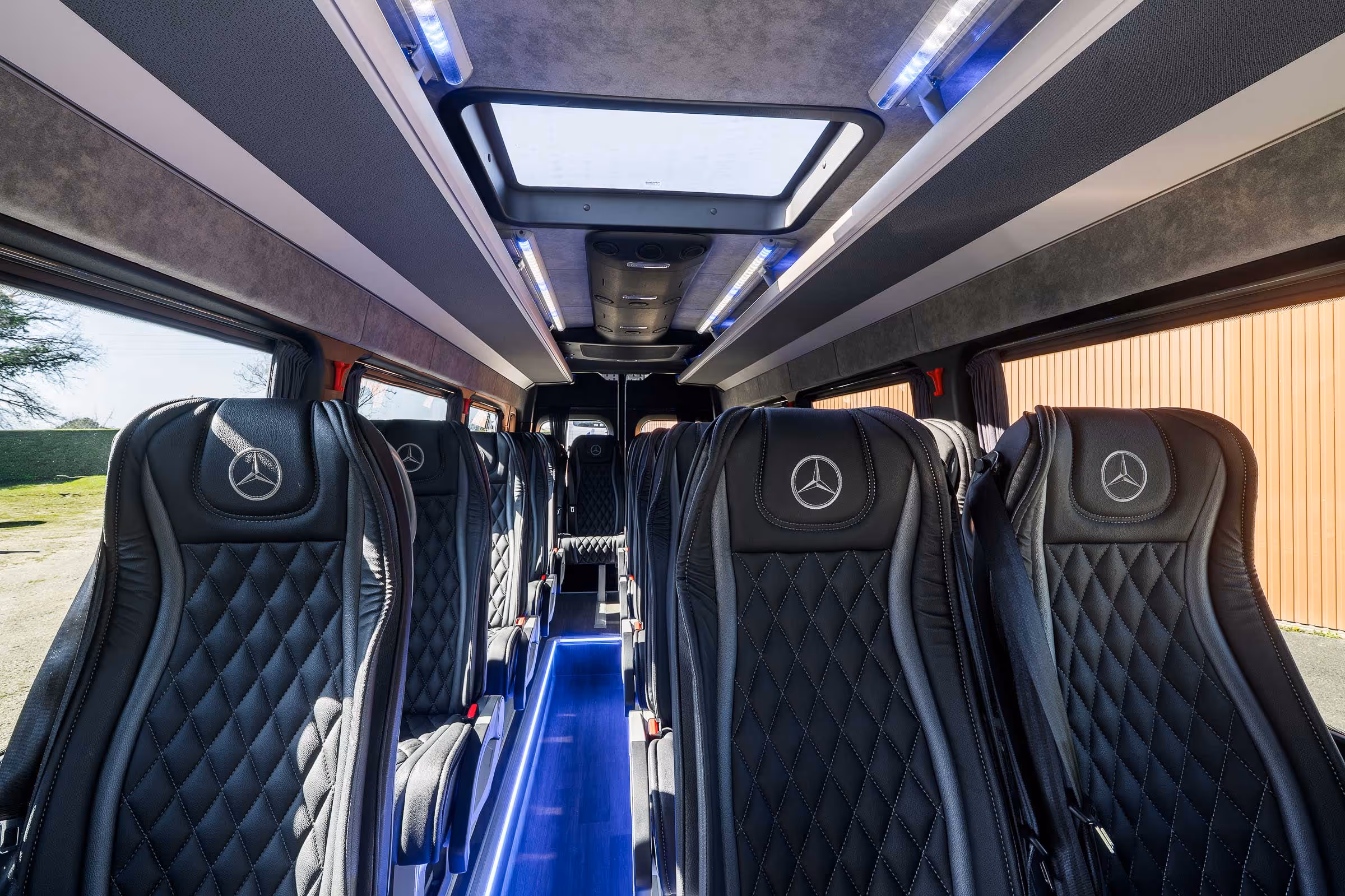 haut de gamme d'un minibus avec des sièges en cuir noir siglés Mercedes-Benz, un éclairage LED bleu au sol et un puits de lumière au plafond.