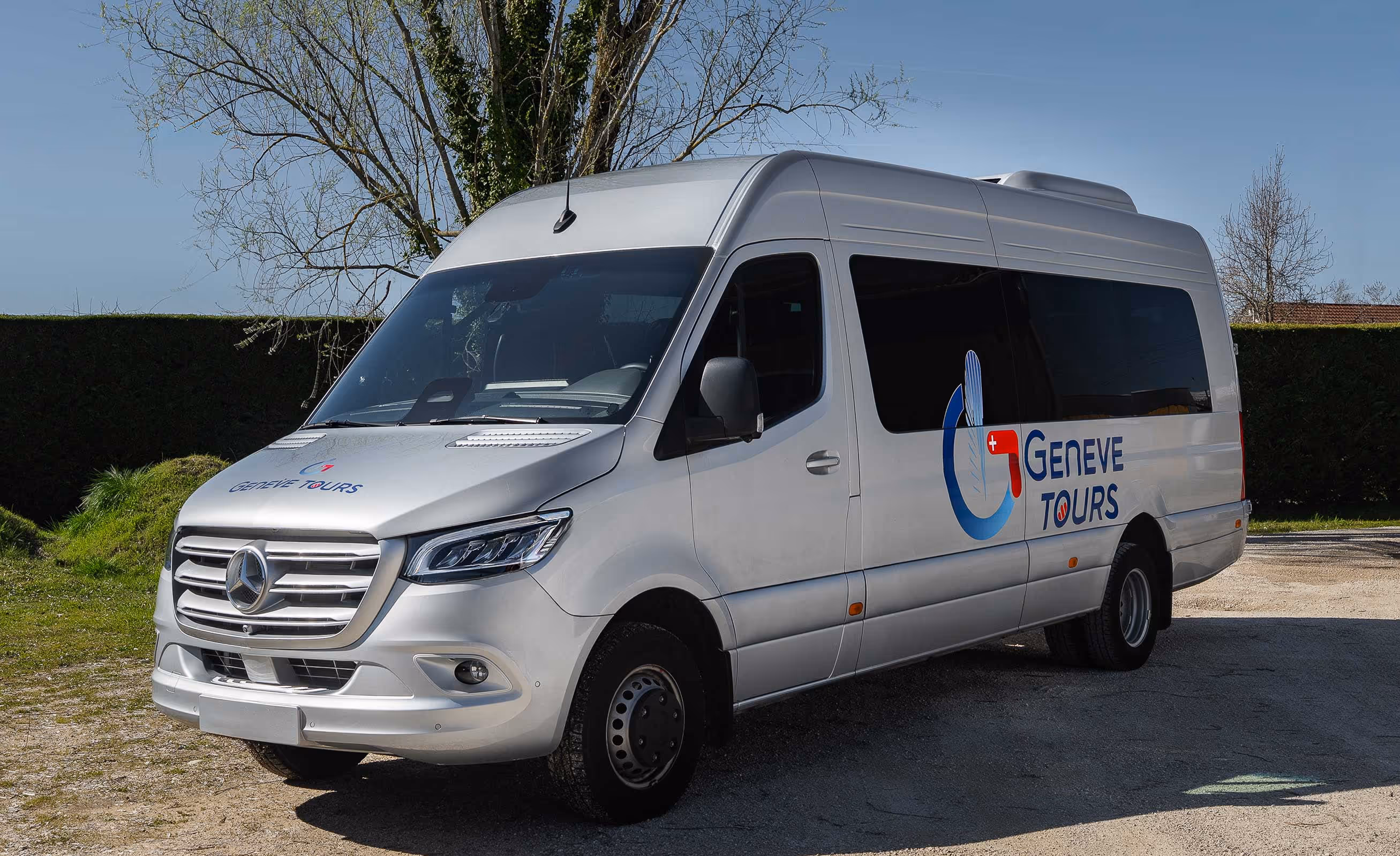 Vue extérieure de trois-quarts d'un minibus Mercedes-Benz gris métallisé arborant le logo "Genève Tours" sur un terrain en gravier.