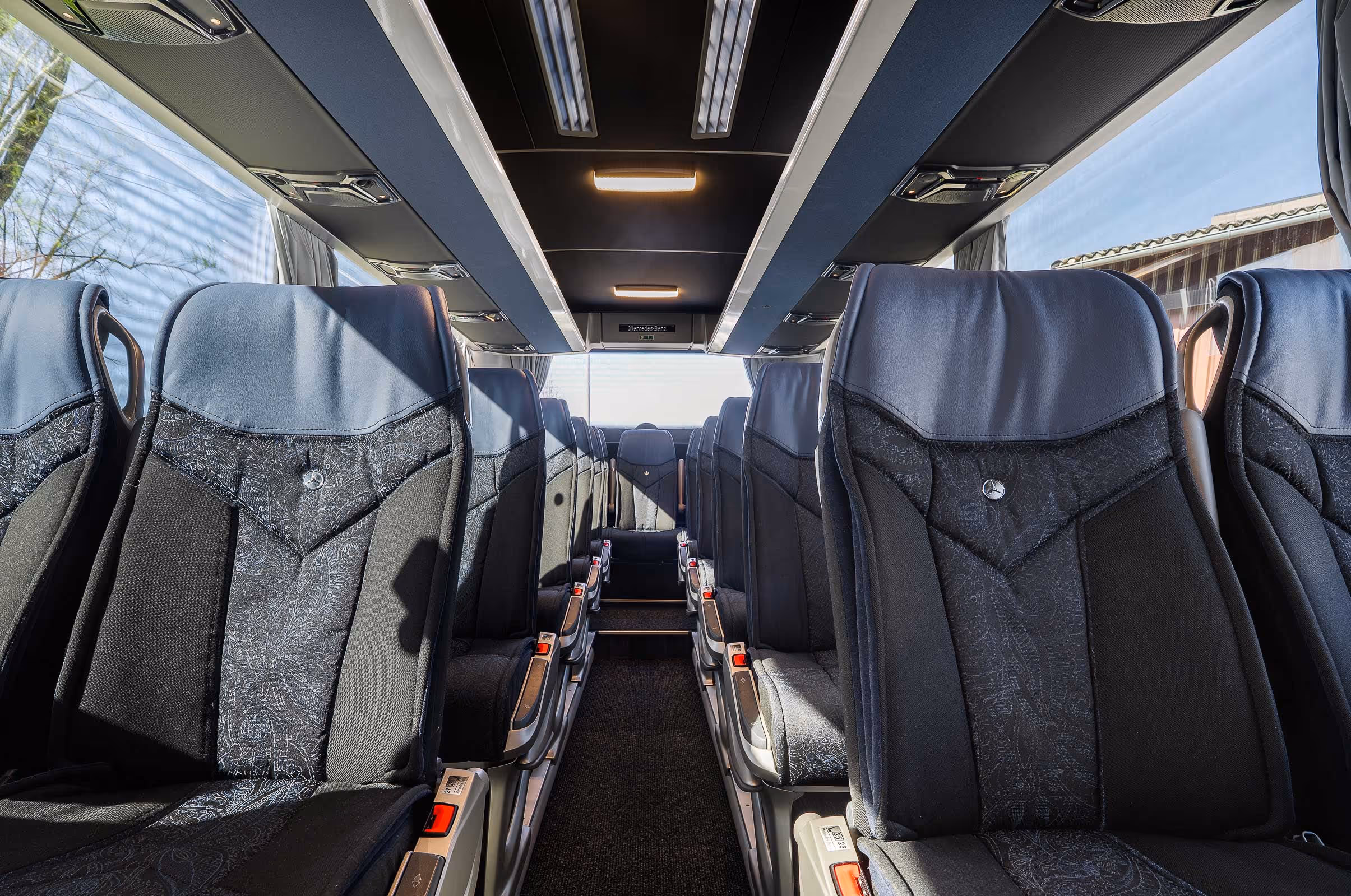 Intérieur d'un autocar avec des sièges en tissu noir et gris, dotés de tablettes individuelles et de l'emblème Mercedes-Benz.