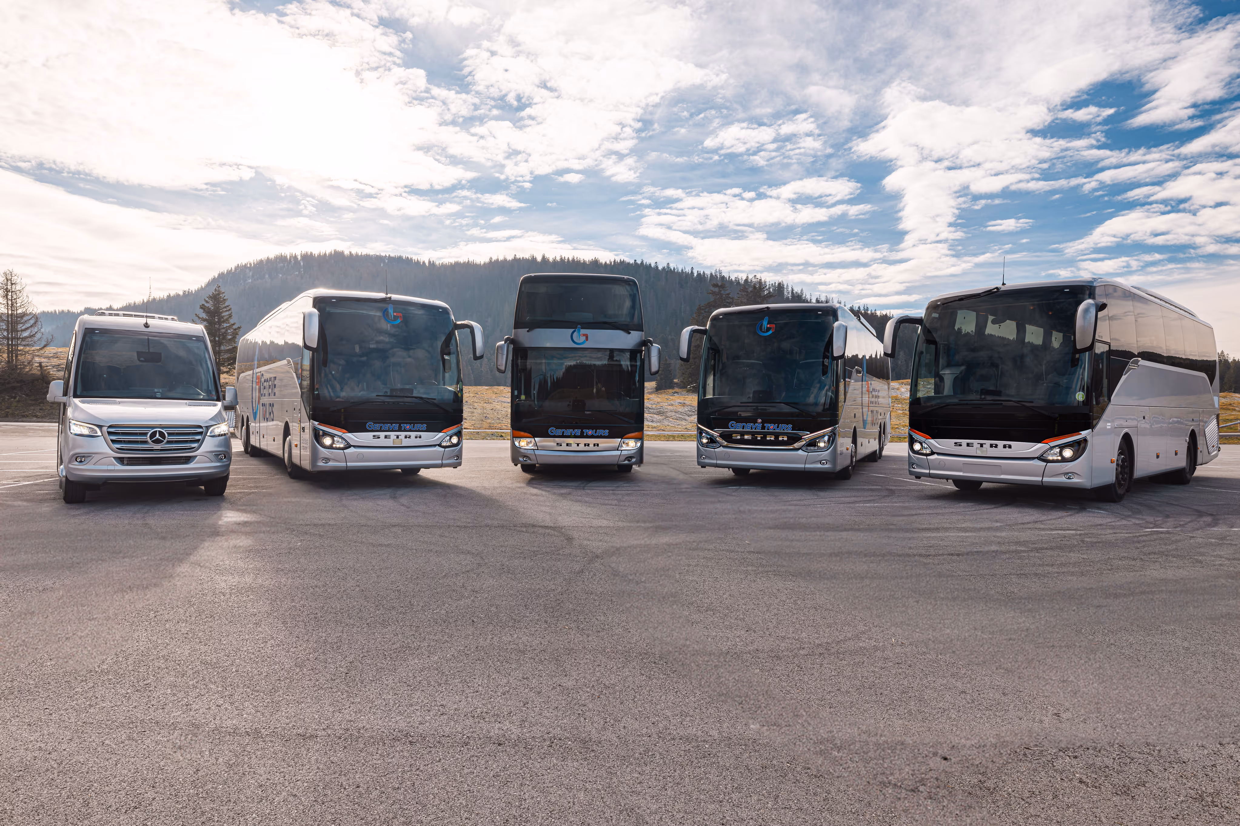 Cinq autocars blancs Mercedes et Setra garés en rangée sur un grand parking avec une forêt et un ciel partiellement nuageux en arrière-plan.