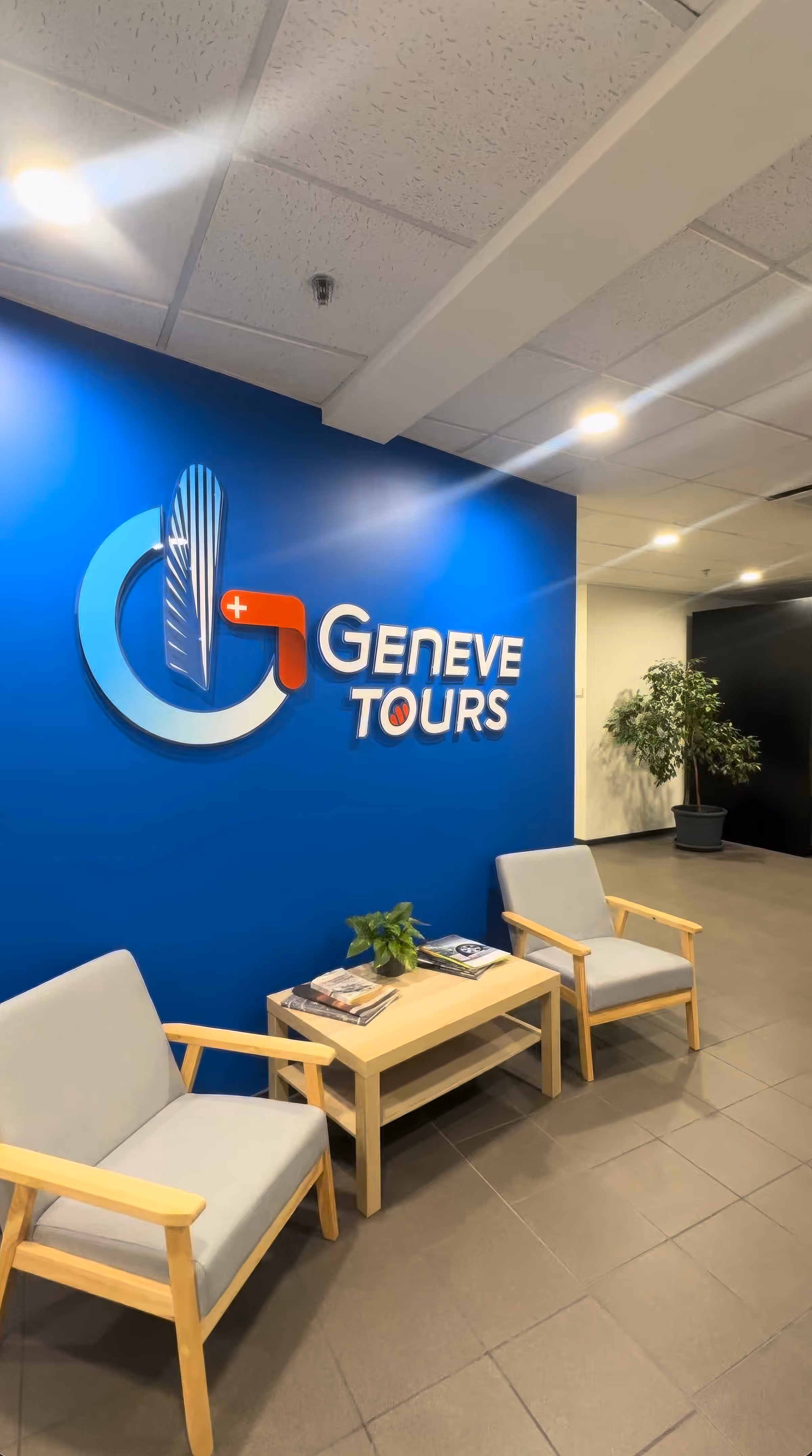 Coin salon avec deux fauteuils en bois clair, une table basse avec des magazines et une plante, devant un mur bleu avec le logo et texte Genève Tours.