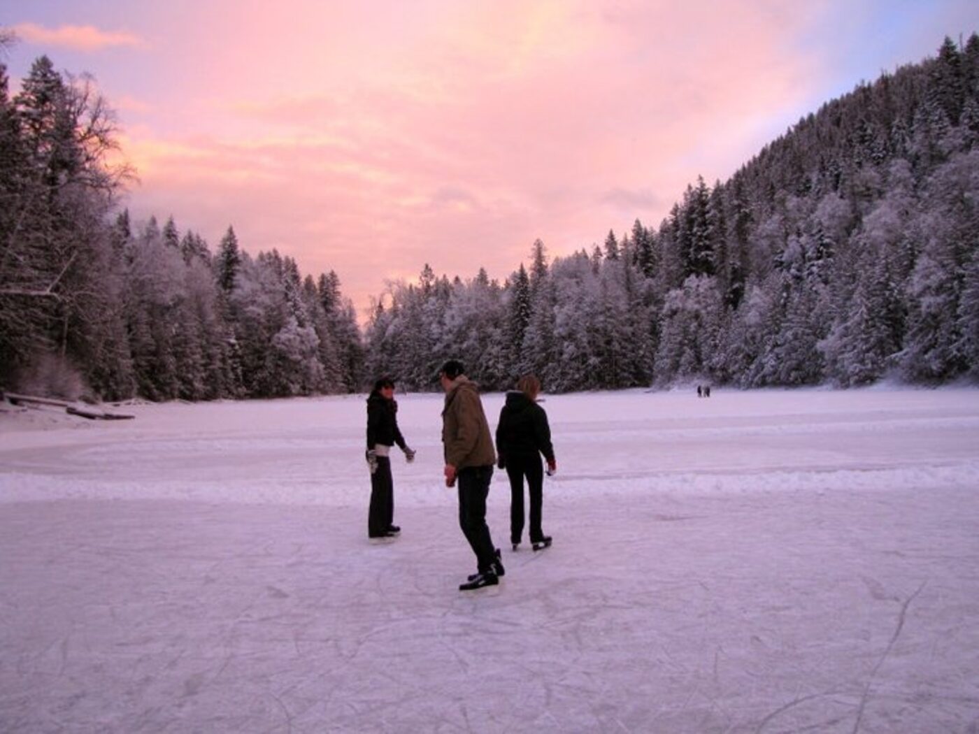 Three people out on the frozen lake under a pink winter sunset, snow covered forest all around