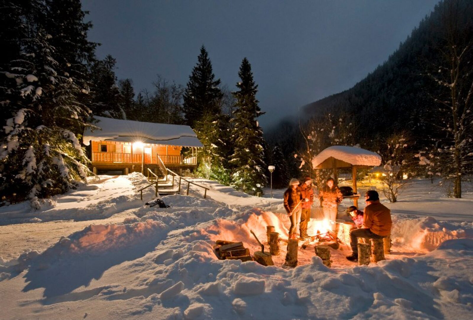 People gathered around an outdoor fire in deep snow at night with a lit cabin behind them