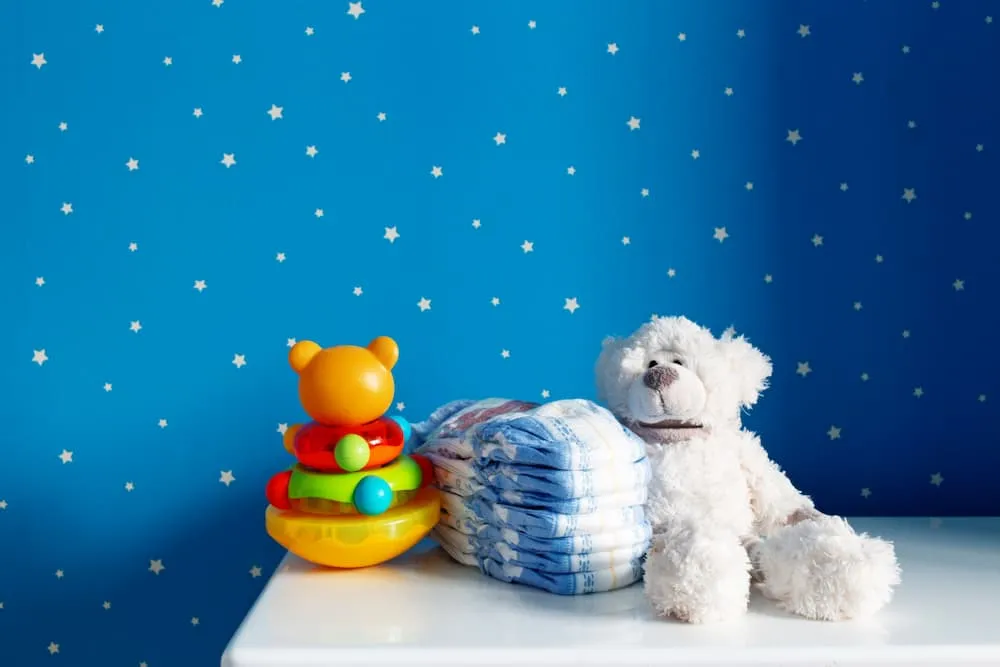 A photo of a teddy bear, diapers and a toy on a changing table in front of a background of blue wallpaper with stars. 