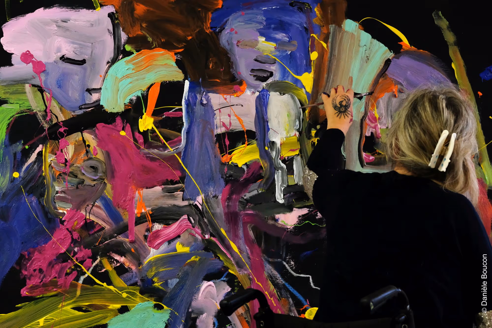 Artiste peignant une grande toile abstraite représentant plusieurs personnages aux couleurs sombres et vives
