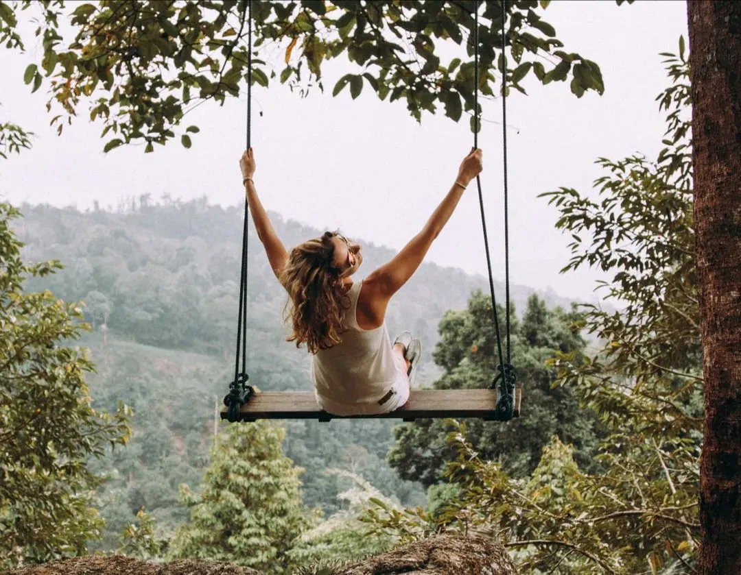 Vrouw op een schommel in de natuur met uitzicht op groene heuvels en bomen.