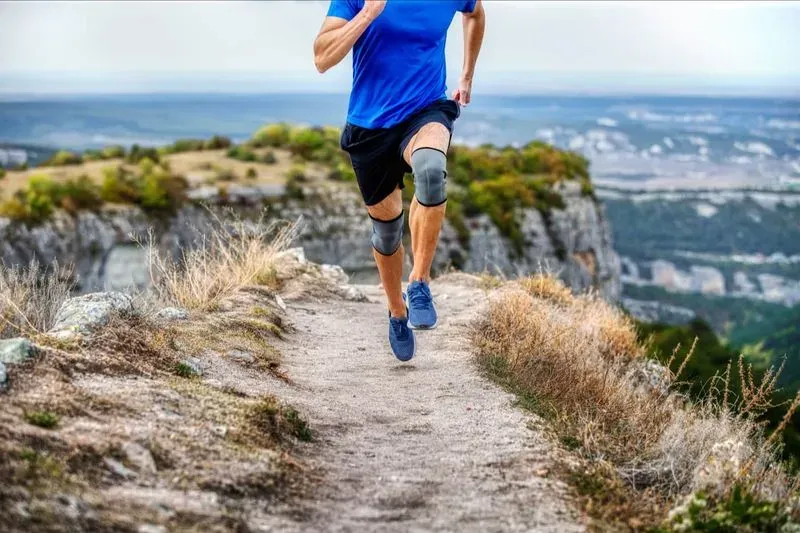 Man met kniebeschermers rent op een smal bergpad met uitzicht op een vallei en rotswanden.