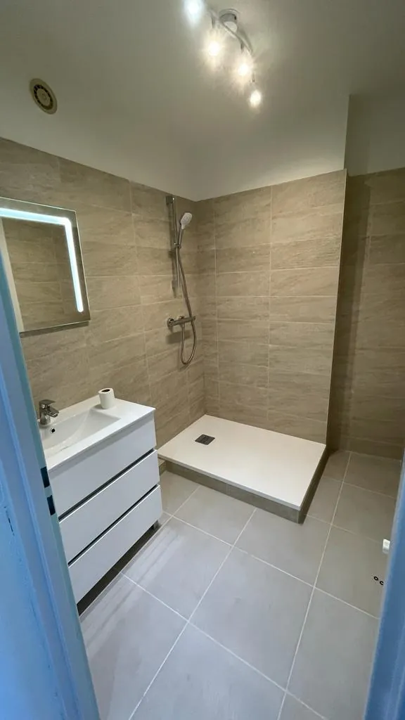 Salle de bain rénovée avec douche à receveur plat, carrelage beige imitation pierre, meuble vasque blanc et miroir LED