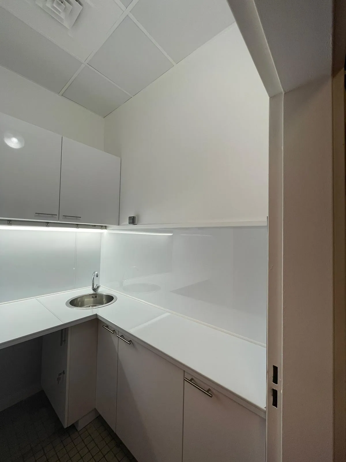 Kitchenette professionnelle en L avec meubles blancs, évier inox rond et éclairage LED sous meuble haut