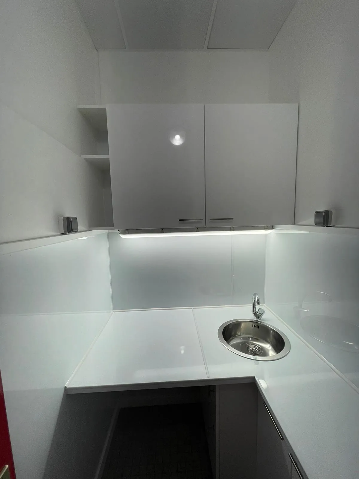 Vue rapprochée de la kitchenette avec plan de travail blanc, évier inox encastré et meubles hauts avec éclairage intégré