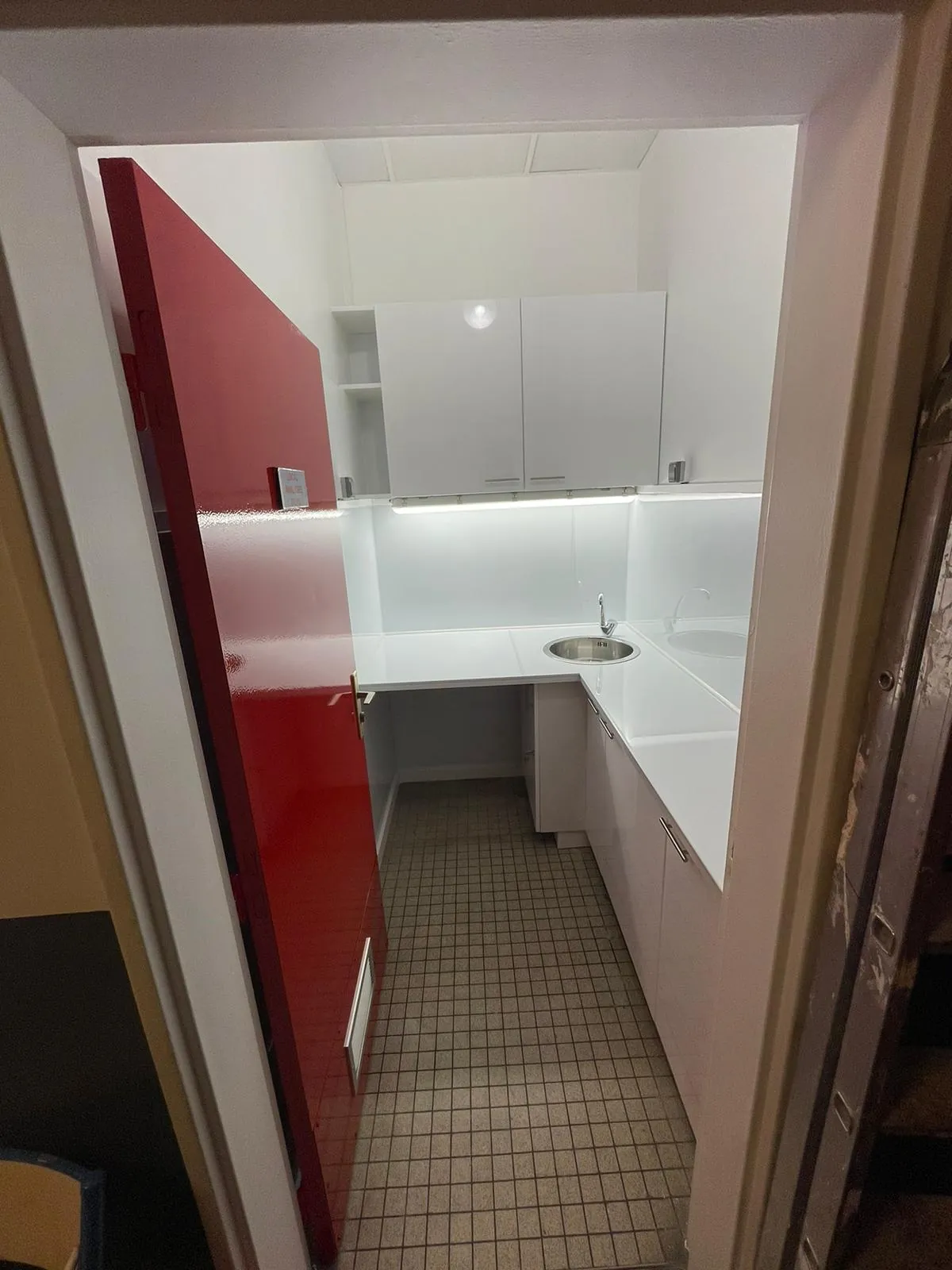Kitchenette neuve avec porte rouge, meubles blancs et évier inox, aménagement de locaux professionnels en régie