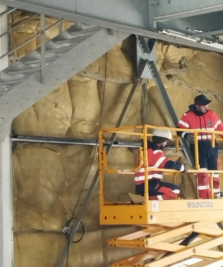 Deux ouvriers du bâtiment en vêtements réfléchissants sur une nacelle à ciseaux jaune à l'intérieur d'un bâtiment en construction.