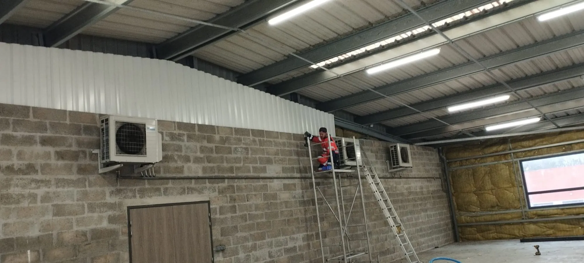 Worker using a handheld circular saw to cut wood on a blue metal scaffold indoors.