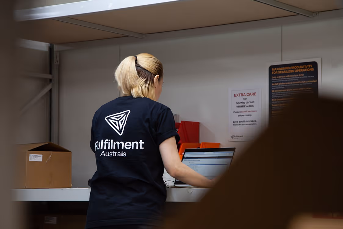 Woman with blonde hair tied in a ponytail wearing a black Fulfilment Australia t-shirt working on a laptop at a shelf in a warehouse.