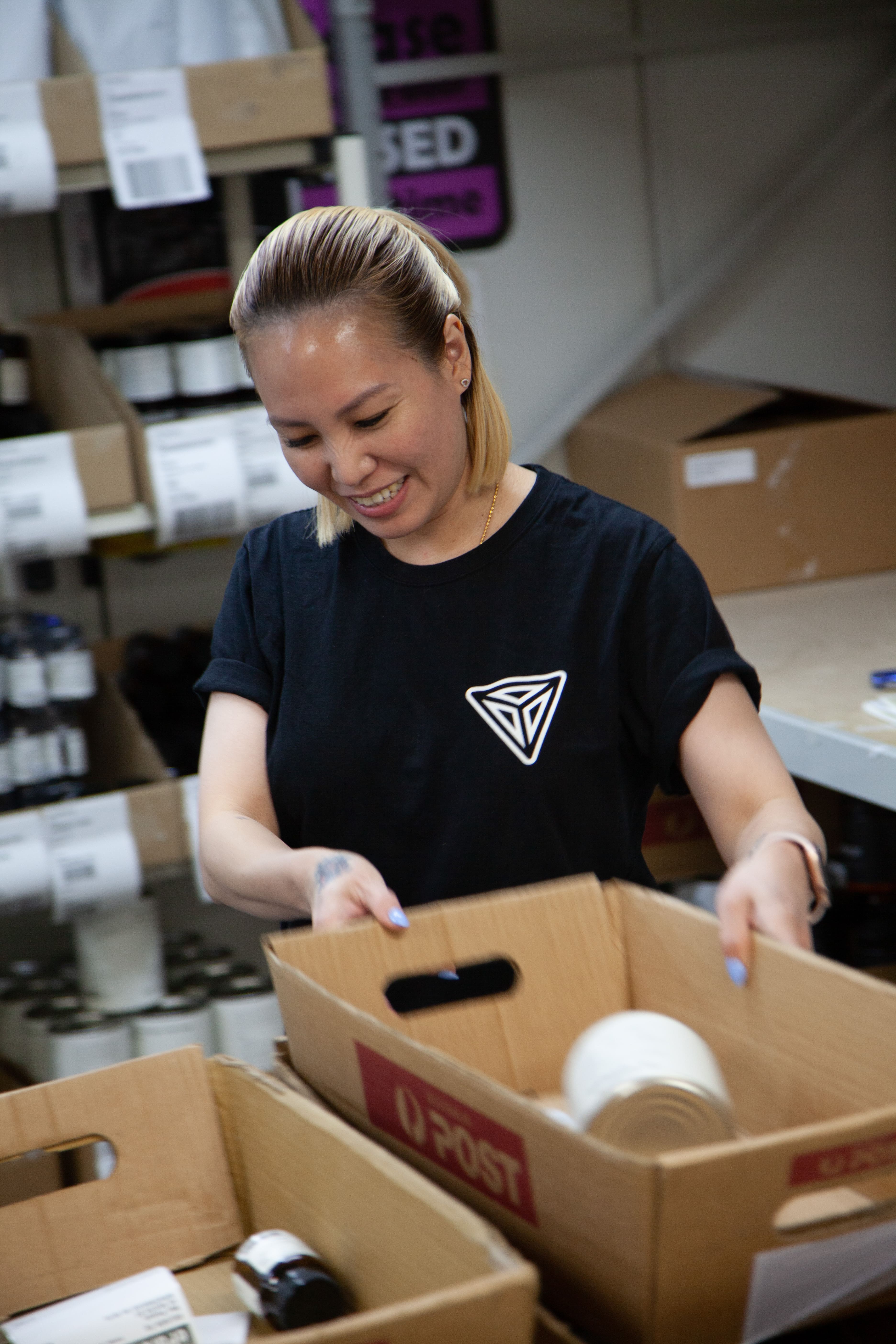 Smiling woman with blonde hair packing or sorting canned goods into cardboard boxes in a storage area.
