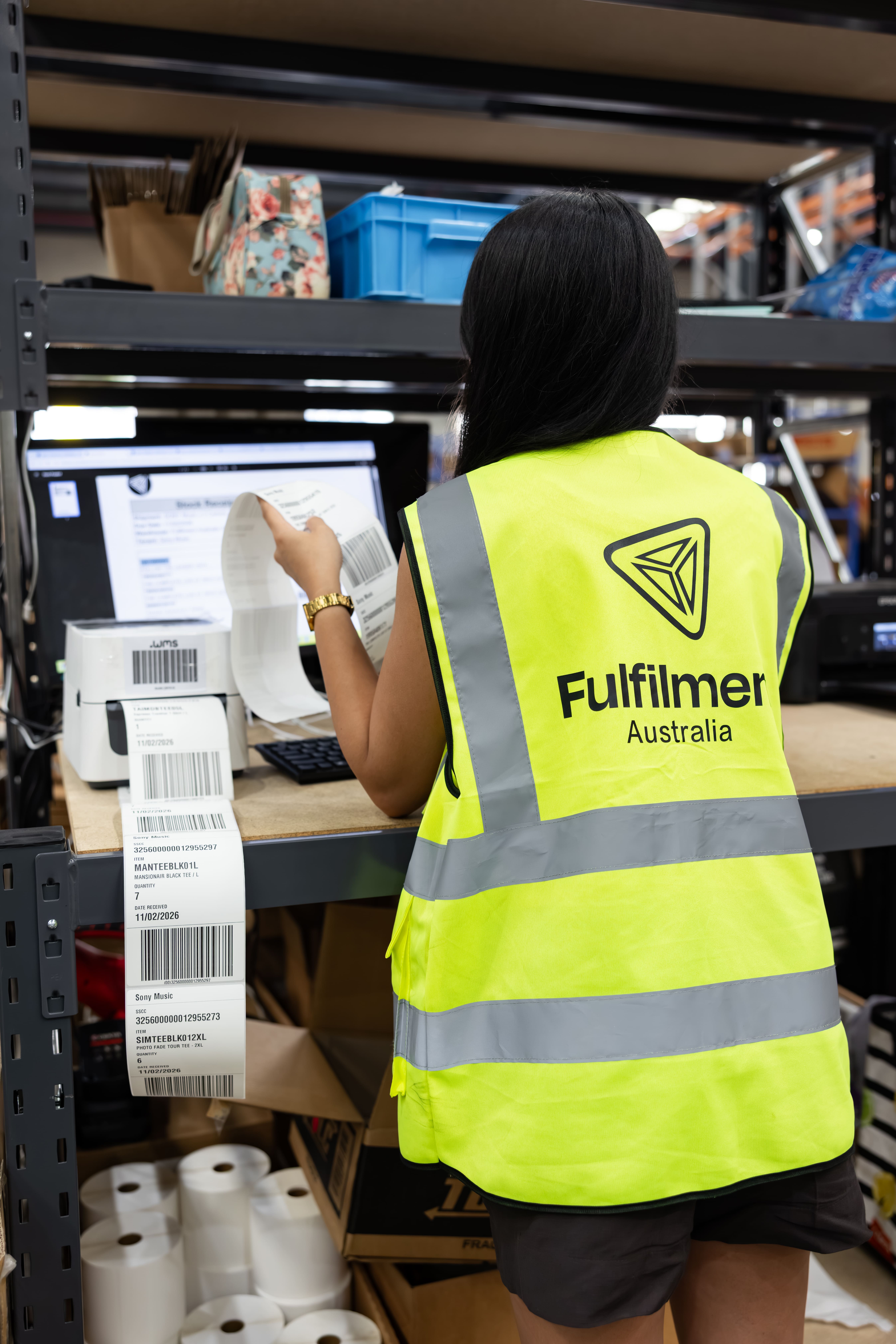 Warehouse worker wearing a yellow high-visibility vest labeled Fulfilmer Australia, holding printed shipping labels in front of a computer and label printer.