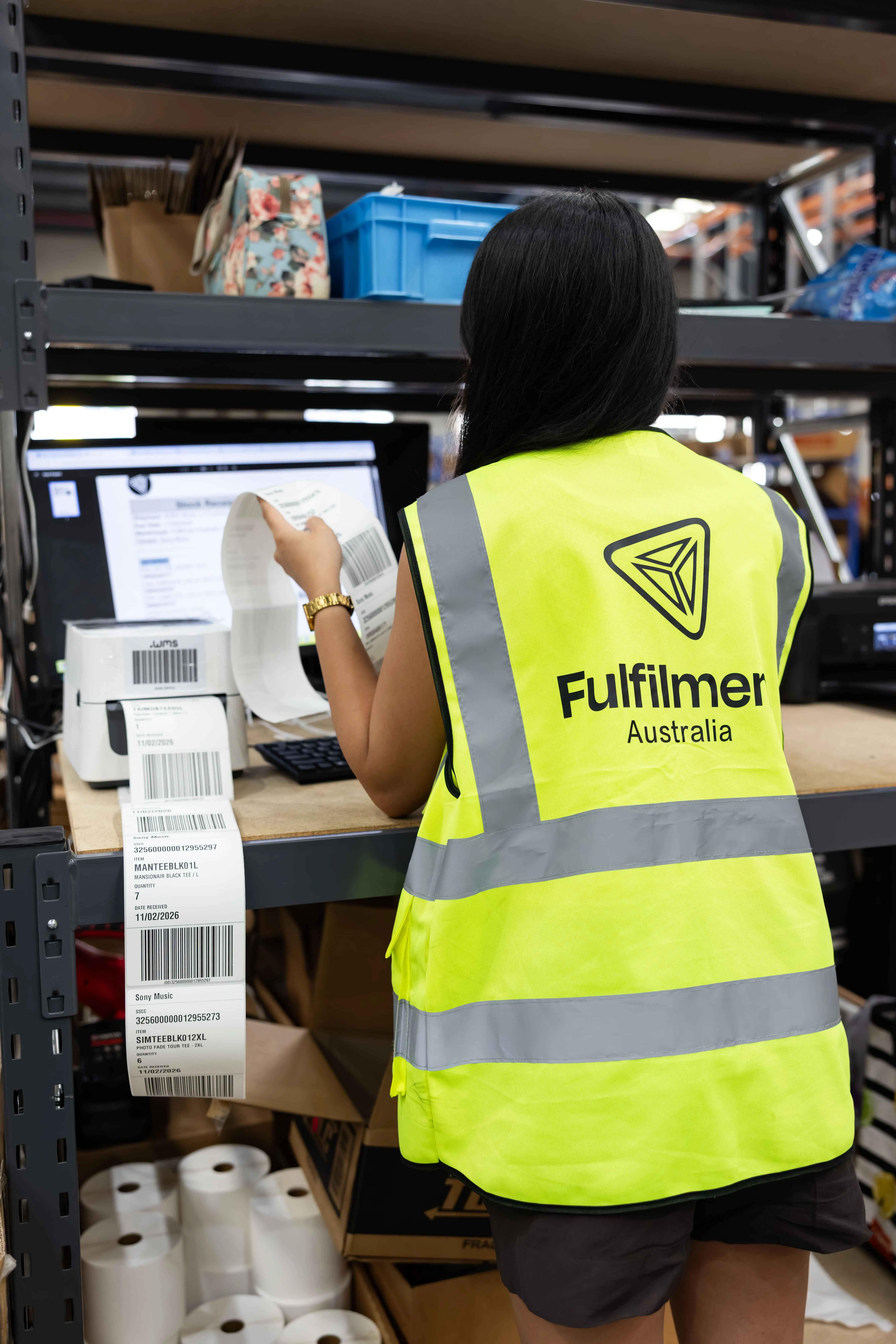 Warehouse worker wearing a yellow high-visibility vest labeled Fulfilment Australia, holding printed shipping labels in front of a computer and label printer.