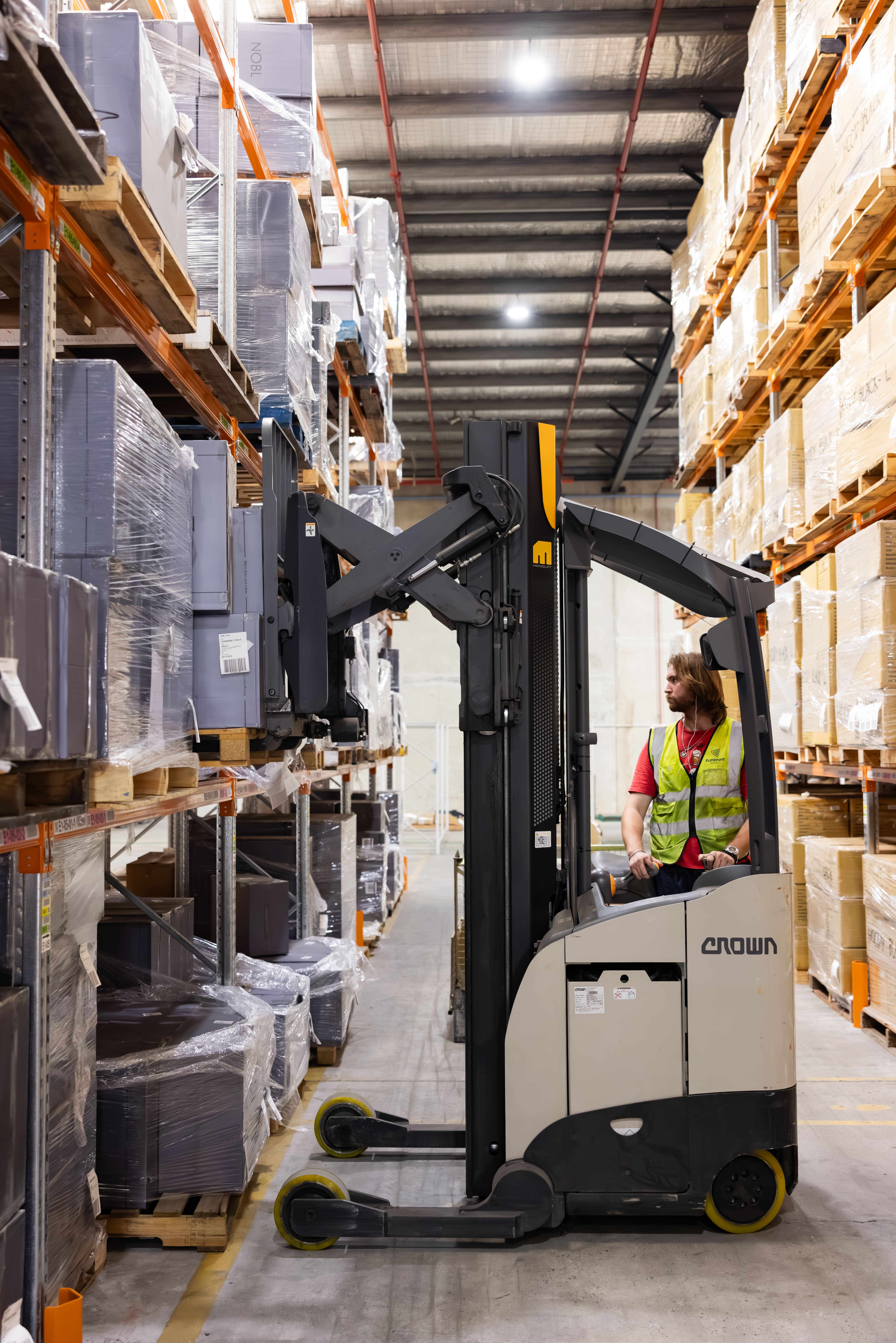 Warehouse worker operating a forklift to lift pallets wrapped in plastic on metal shelves.