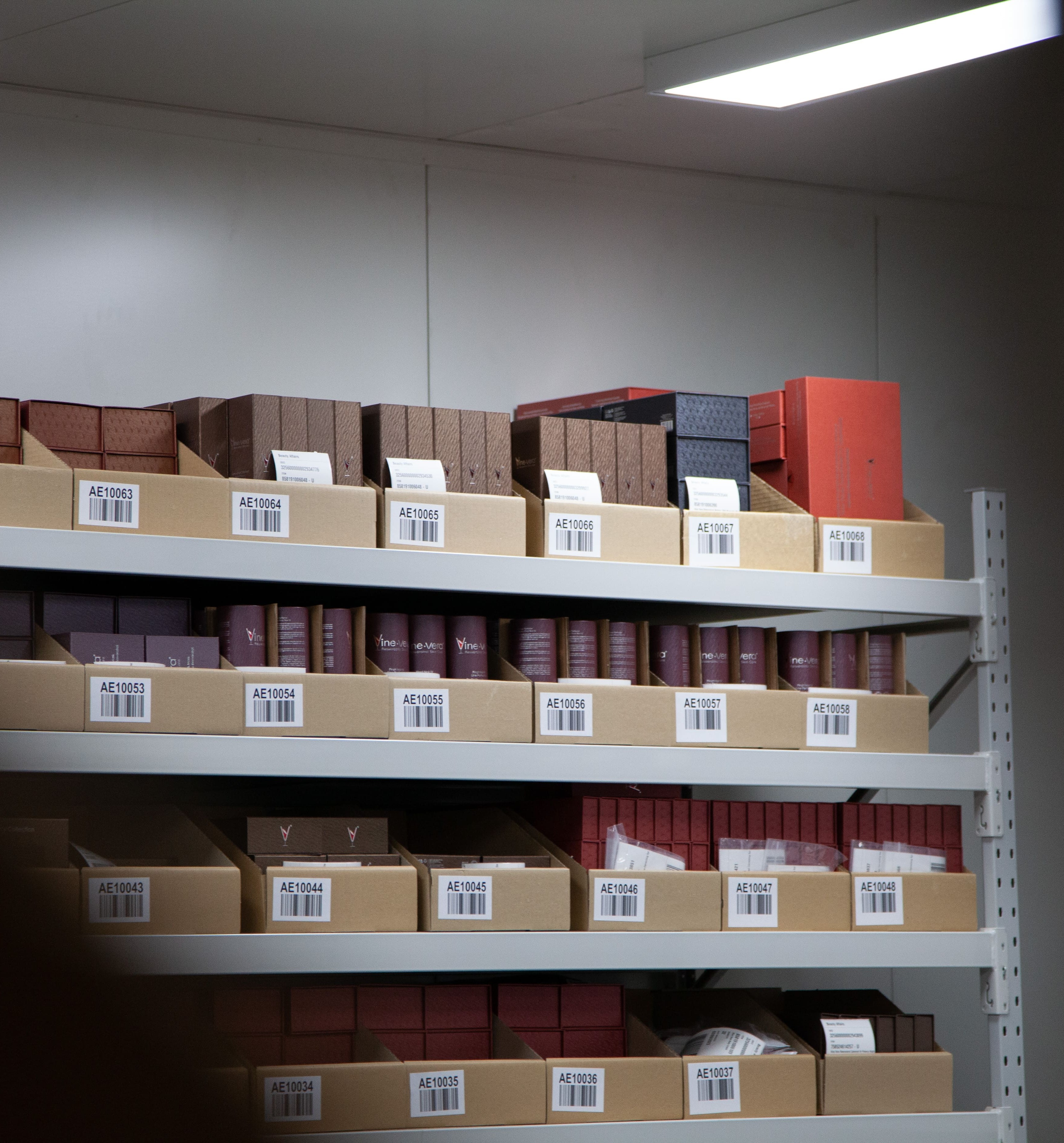 Metal shelving units with cardboard boxes holding various red and brown packaged items, each labeled with barcodes and codes like AE10063 to AE10068.