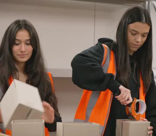 Two women in orange safety vests packing and sealing cardboard boxes in an indoor setting.