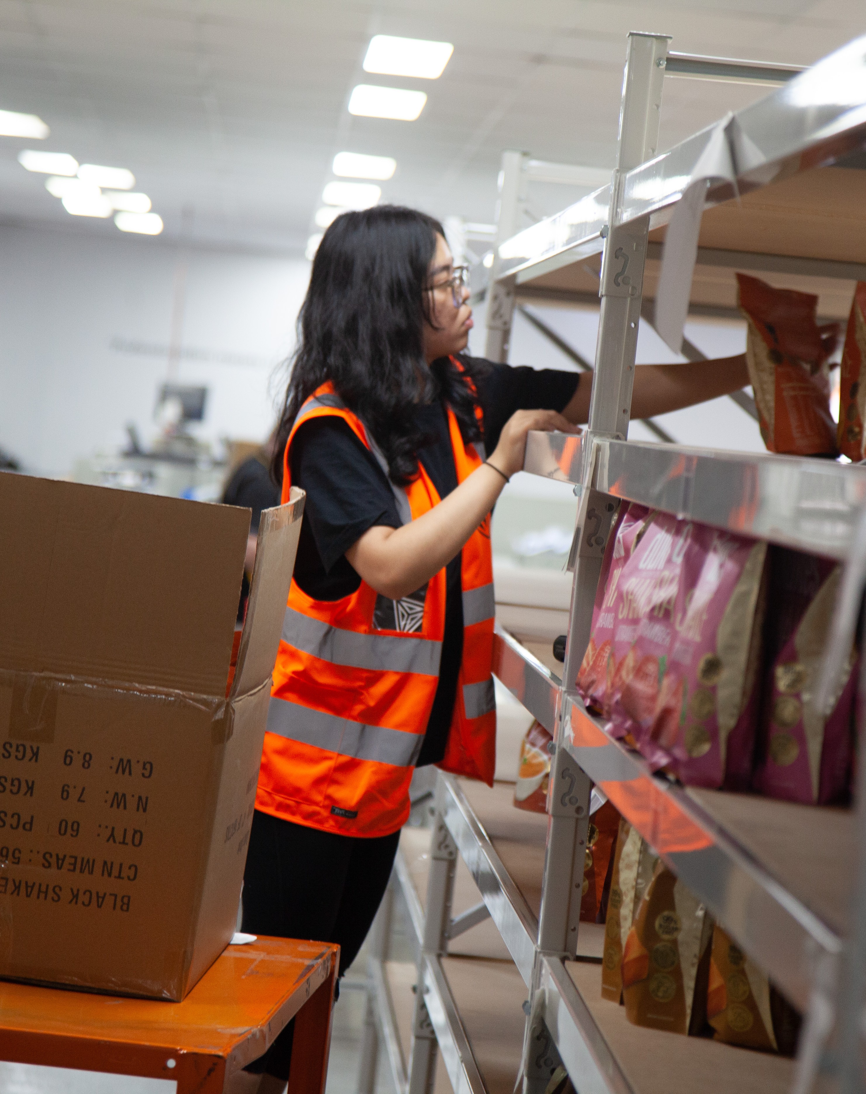Warehouse worker wearing an orange safety vest organizing packaged products on metal shelves.