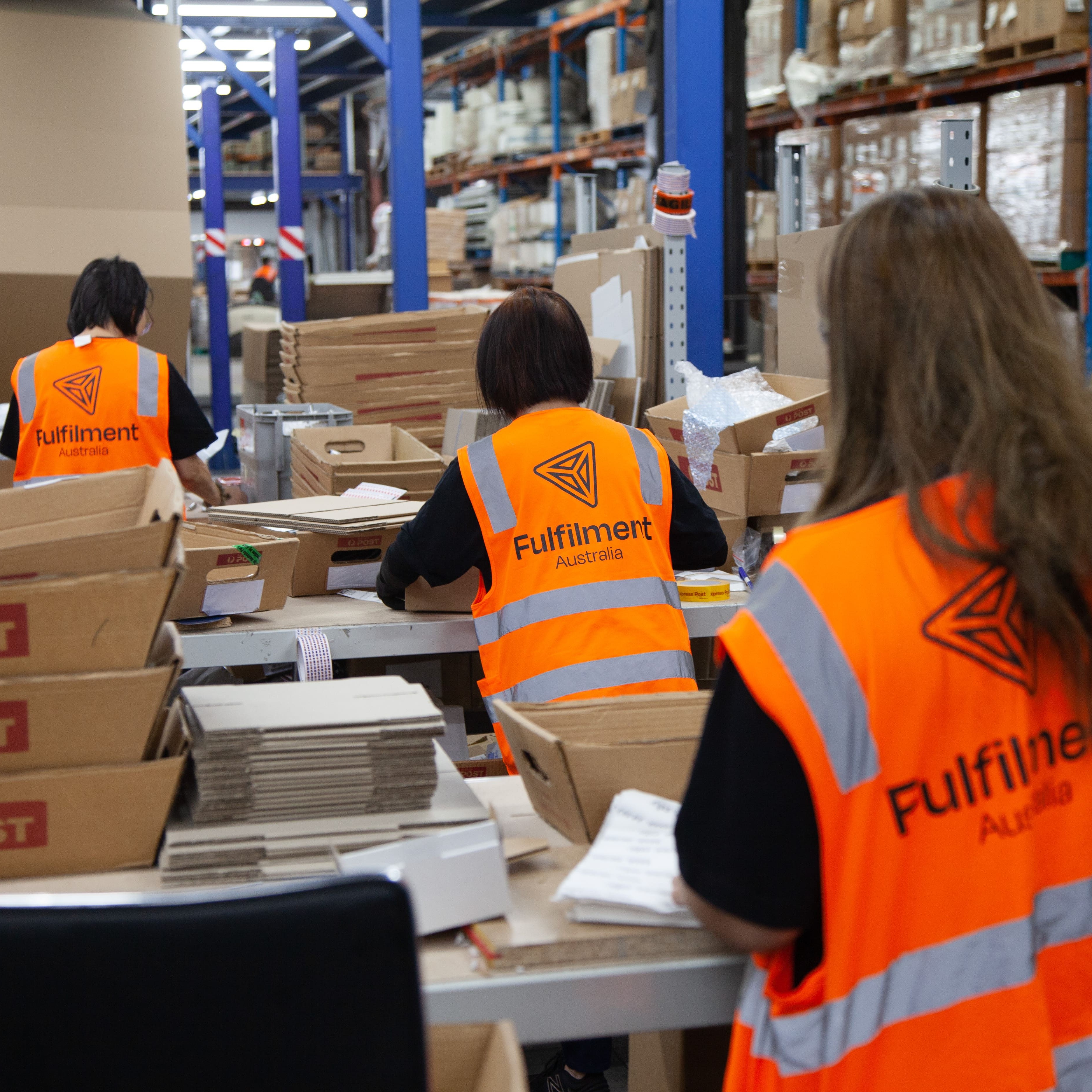Three workers wearing orange Fulfilment Australia safety vests packing boxes in a busy warehouse.