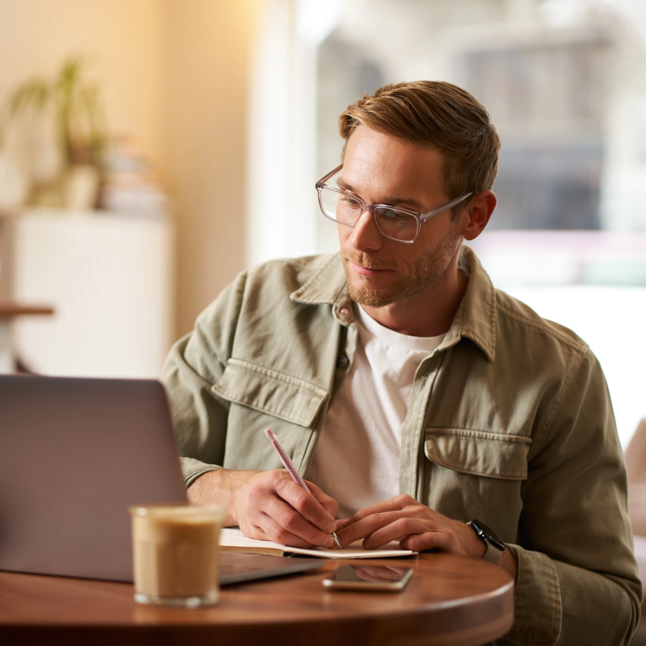 Man with glasses sitting at a table, writing in a notebook with a laptop and coffee nearby.