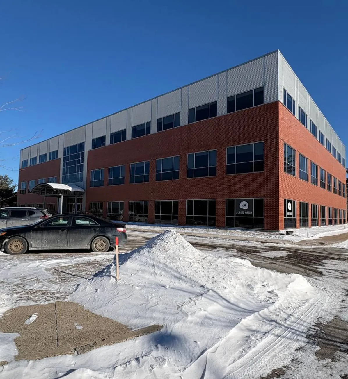 Three-story commercial building with brick and light gray siding, surrounded by snow and parked cars under a clear blue sky.