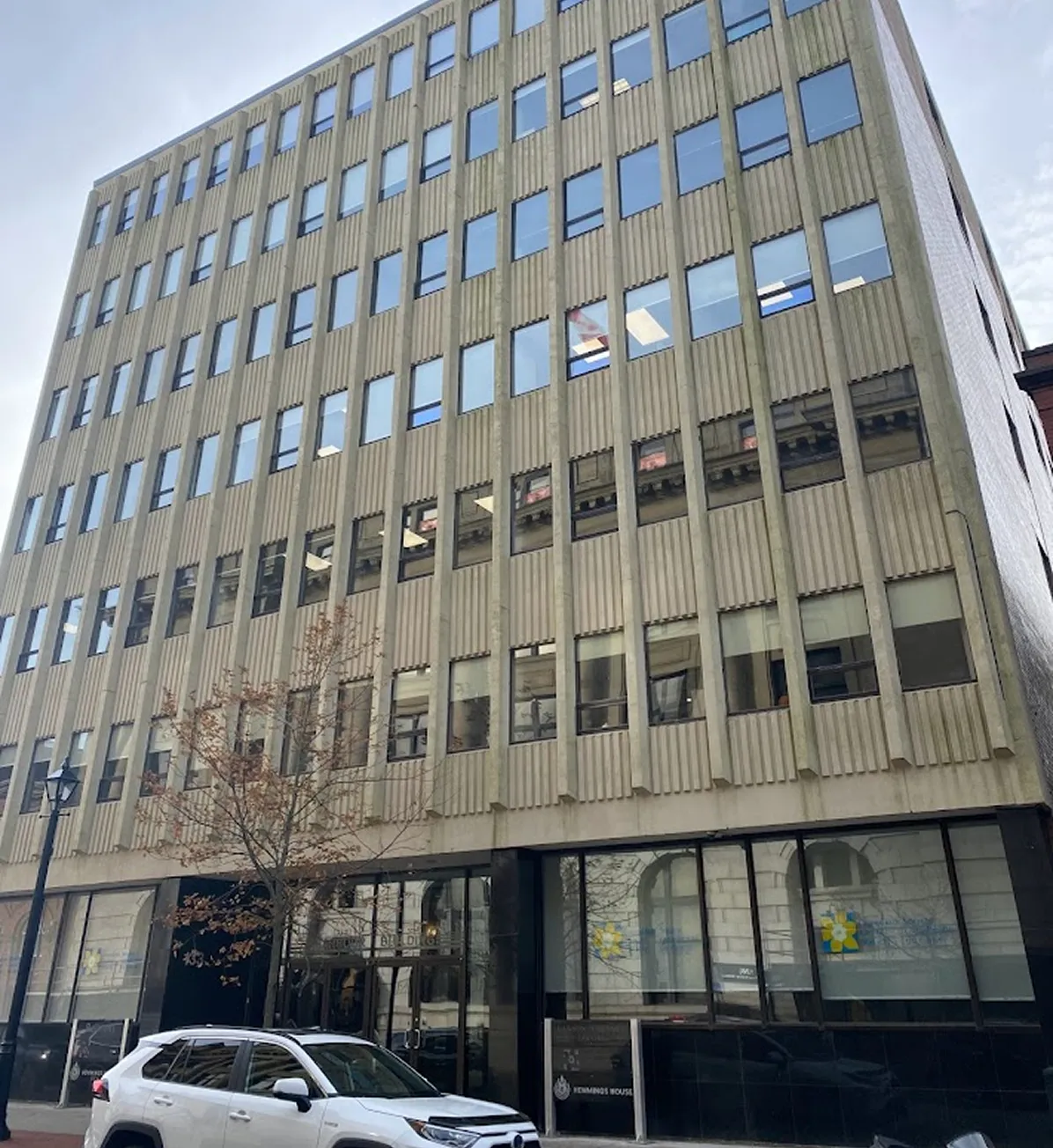 Multi-story office building with vertical concrete panels and many windows, a white car parked in front.