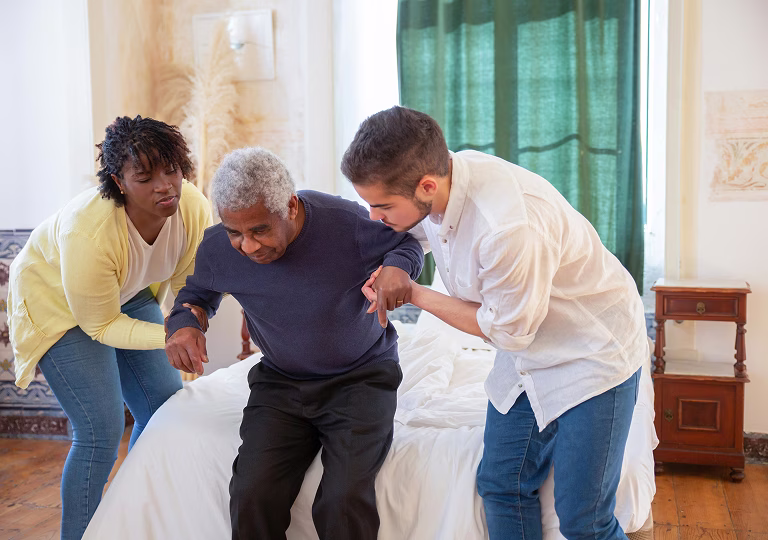 Deux adultes aident un homme âgé à se lever d'un lit dans une chambre lumineuse.