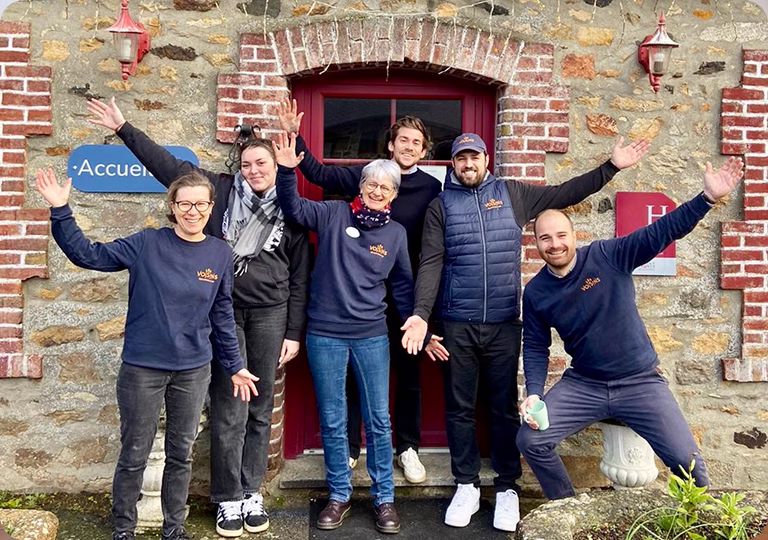 Groupe de six personnes souriantes faisant signe devant une porte rouge encadrée de pierres et briques, devant un mur en pierre avec des panneaux Accueil et H.