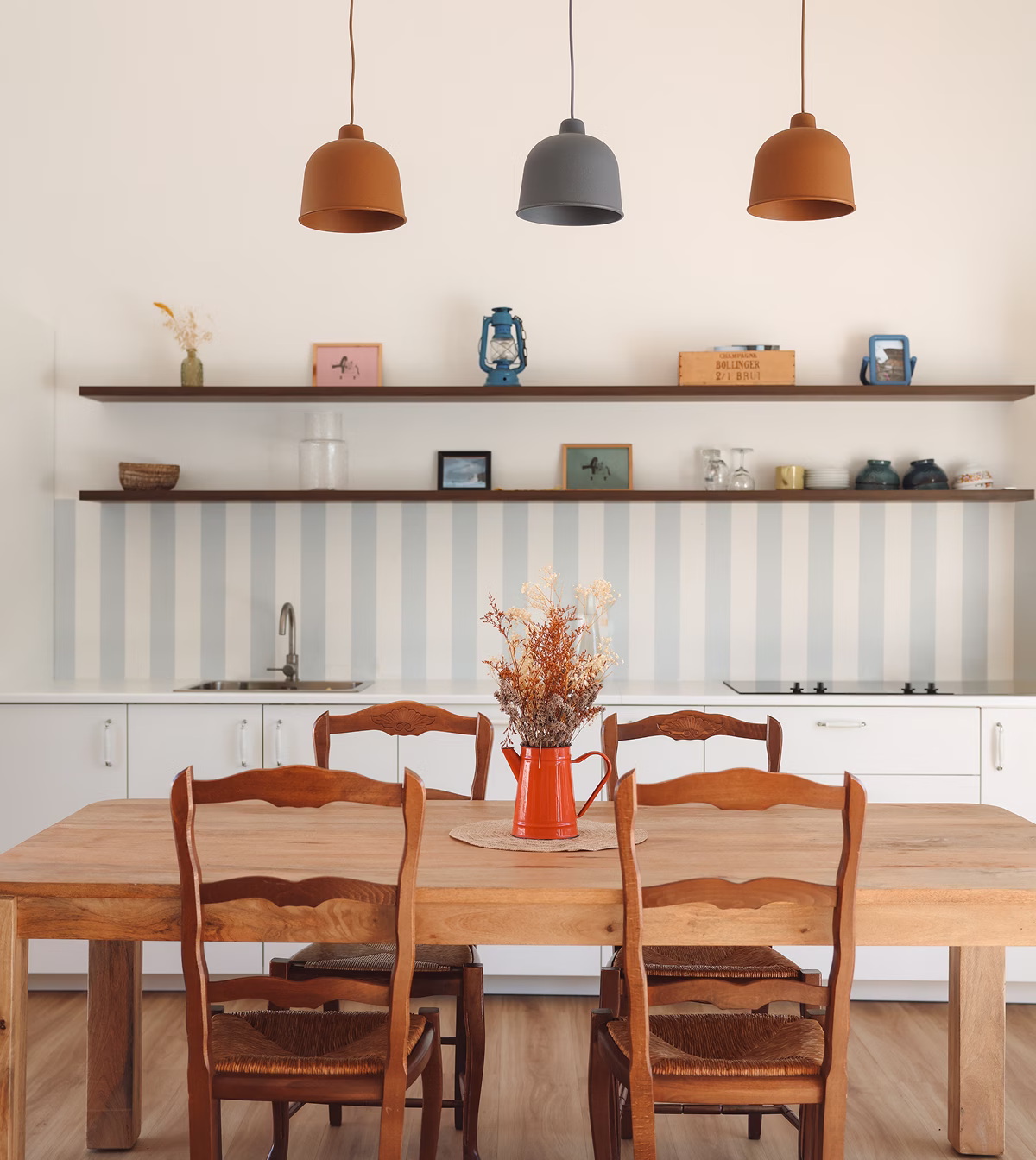 Cuisine moderne avec table en bois, quatre chaises, suspension marron et grise, et bouquet de fleurs séchées dans un vase orangé.