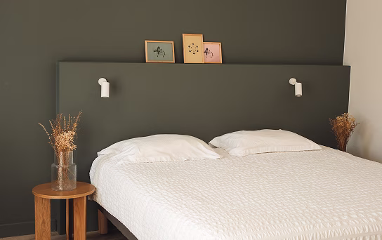 Chambre avec un grand lit double à tête de lit grise, deux lampes murales blanches, deux cadres sur la tête de lit, et une table ronde en bois avec un vase de fleurs séchées.