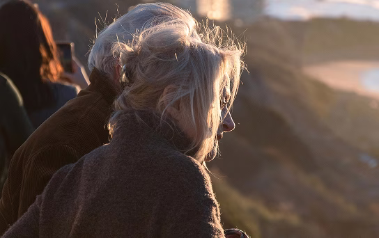 Deux personnes âgées contemplant un paysage vallonné au coucher du soleil.