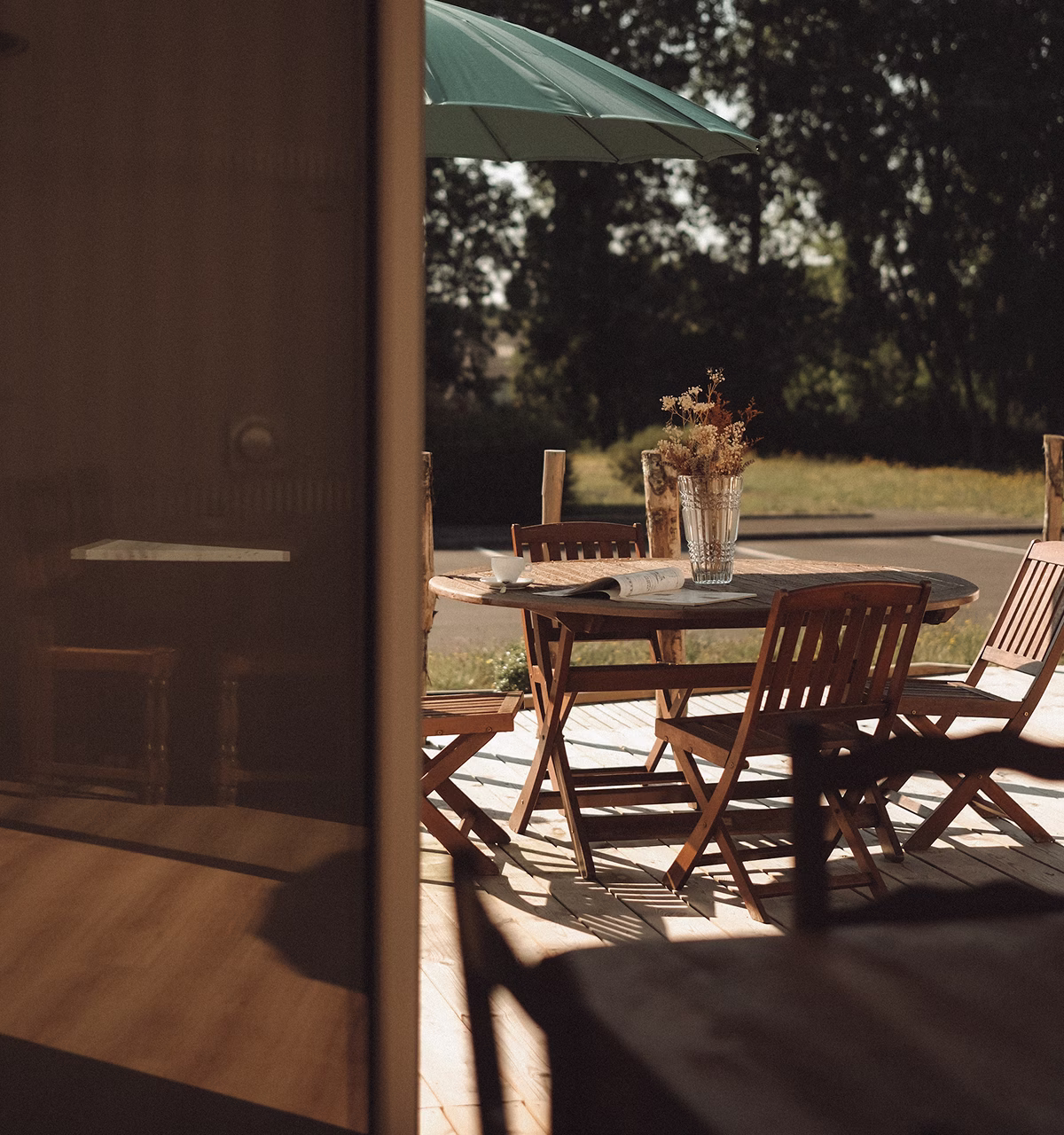 Table en bois avec des chaises sur une terrasse ensoleillée, décorée d'un vase de fleurs séchées et d'une tasse.