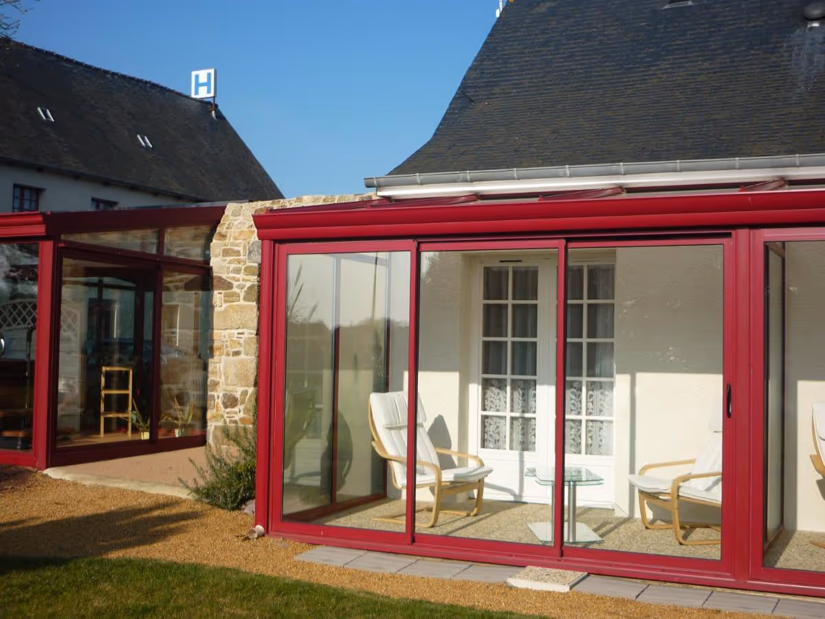 Véranda en verre avec structure rouge devant une maison à toit en ardoise, avec deux fauteuils blancs et une petite table en verre à l'intérieur.