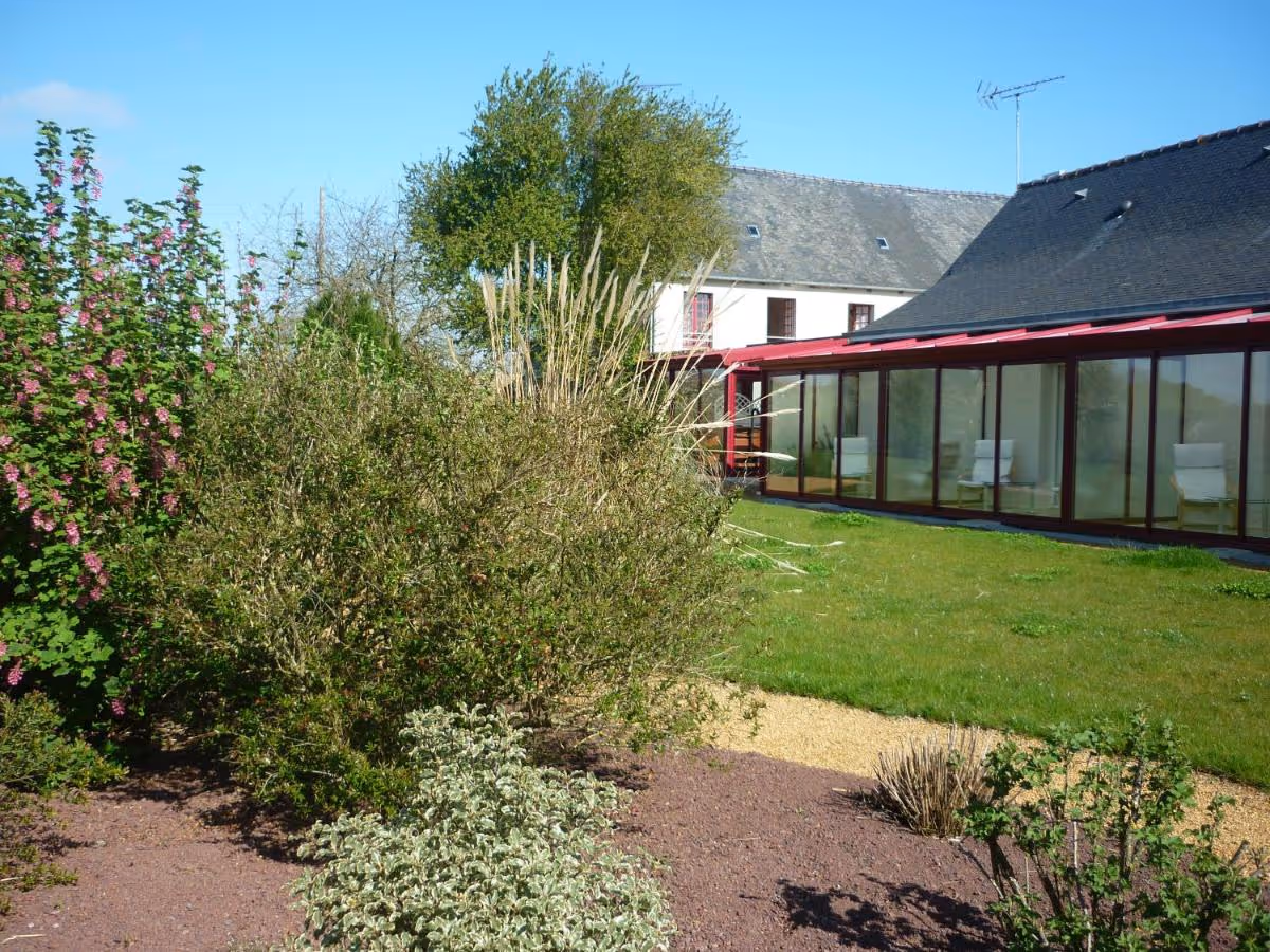 Maison avec une véranda vitrée à structure rouge, jardin avec pelouse et diverses plantes sous un ciel bleu clair.