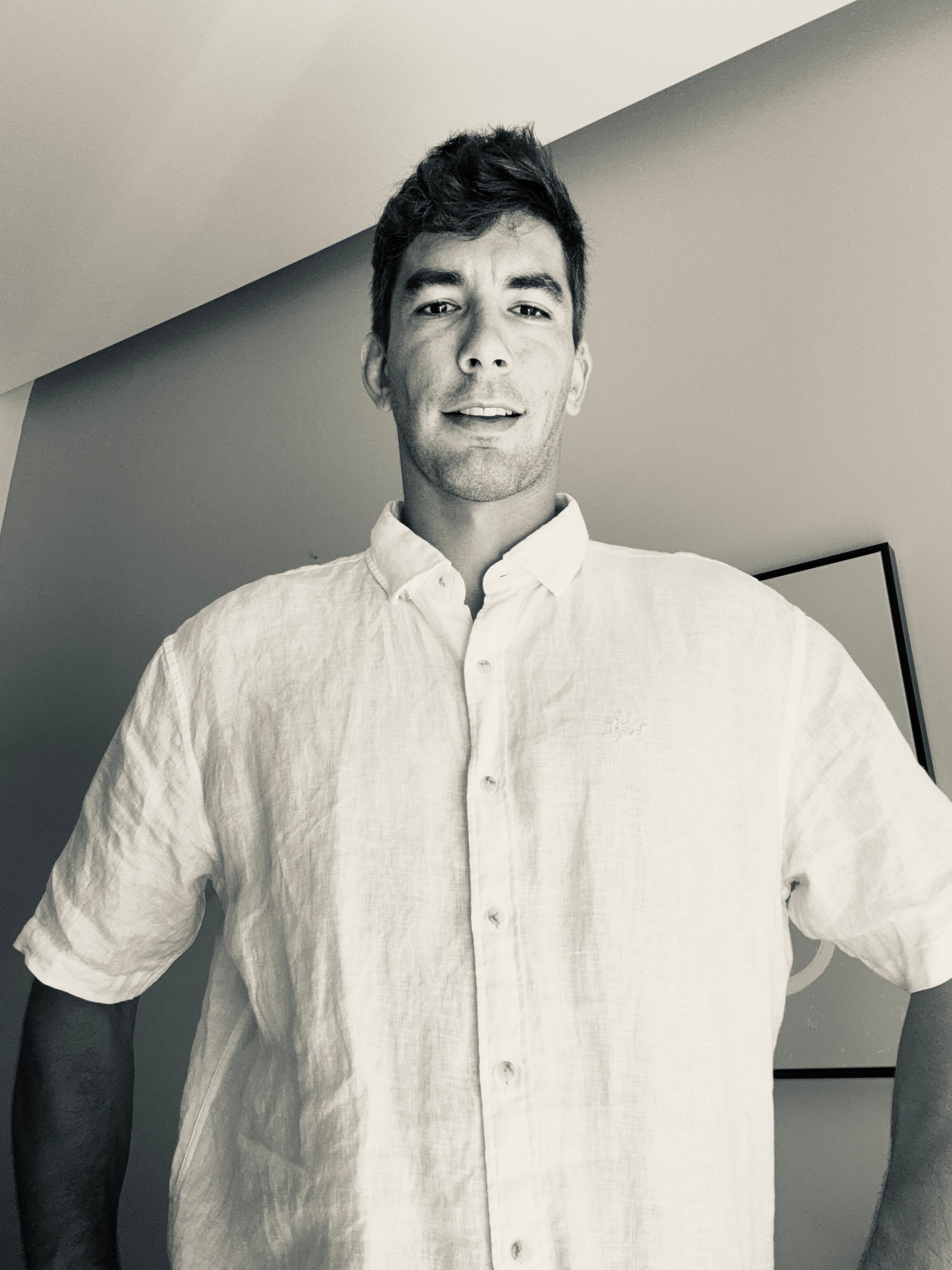 Retrato en blanco y negro de un hombre joven de pie con camisa blanca de manga corta, mirando a la cámara. Es Sebastian Valenzuela consultor de Intelegencia Artificial.