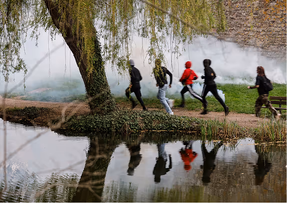 Groupe de cinq personnes courant sur un sentier près d'un arbre au bord d'un plan d'eau, avec de la fumée en arrière-plan.