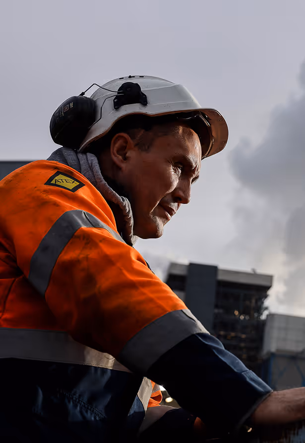 Homme portant un casque de sécurité blanc et un vêtement orange haute visibilité avec un patch ATEX, regardant vers la droite avec un bâtiment industriel en arrière-plan.