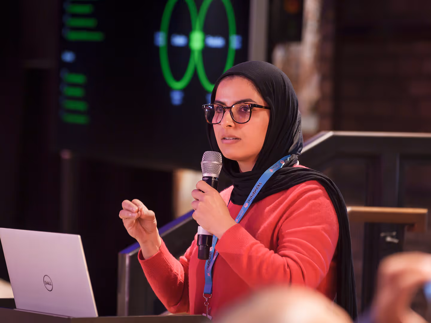 MLOps speaker in a headscarf presenting with a microphone beside a laptop and projected diagram