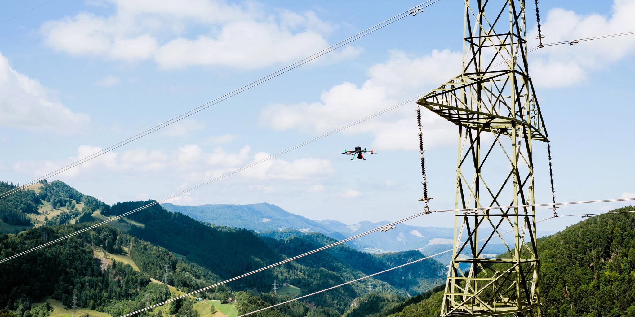 Inspection drone flying alongside a high-voltage transmission tower over a forested valley