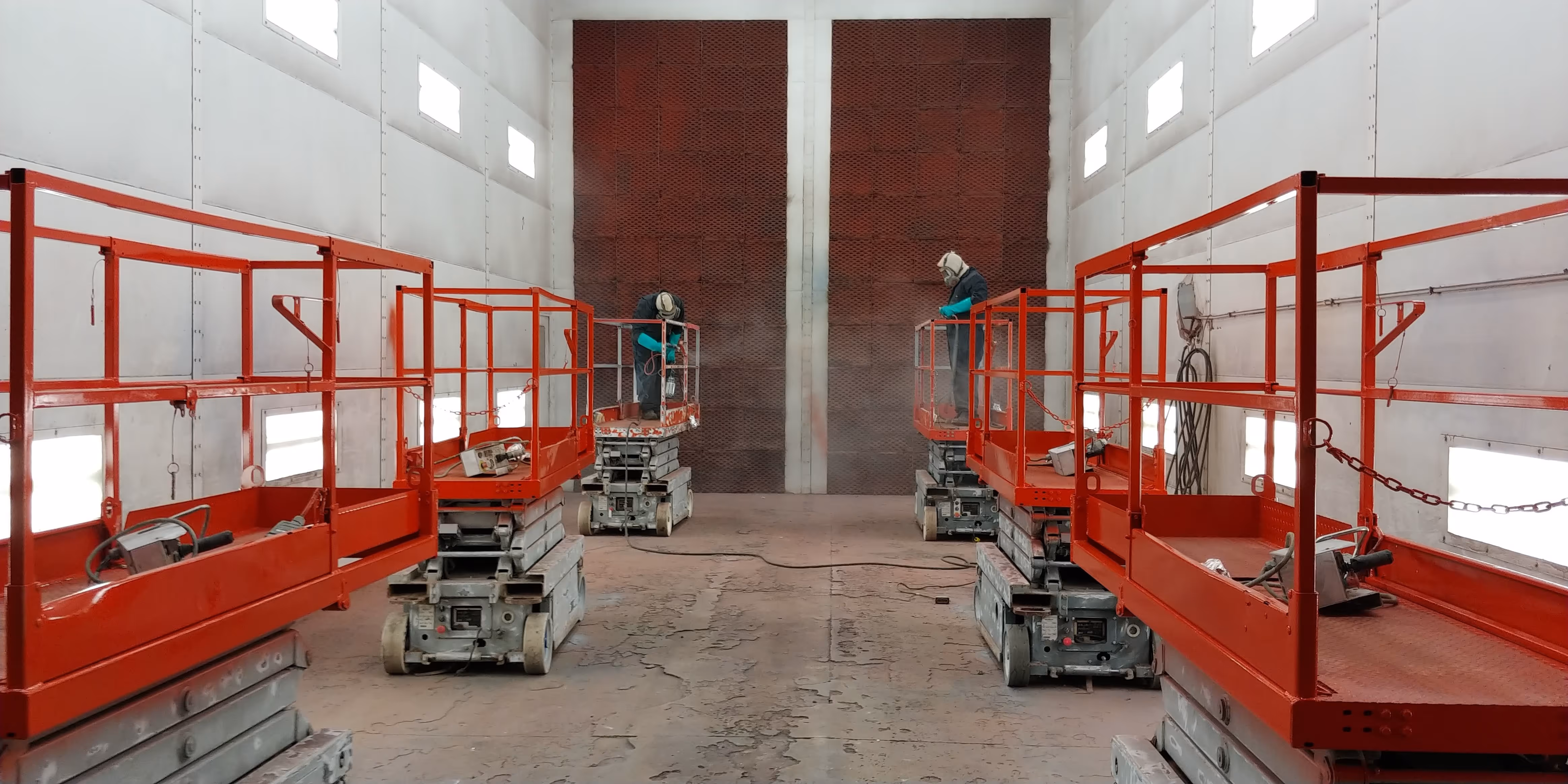 two painters painting scissor lifts the color red inside the paint booth