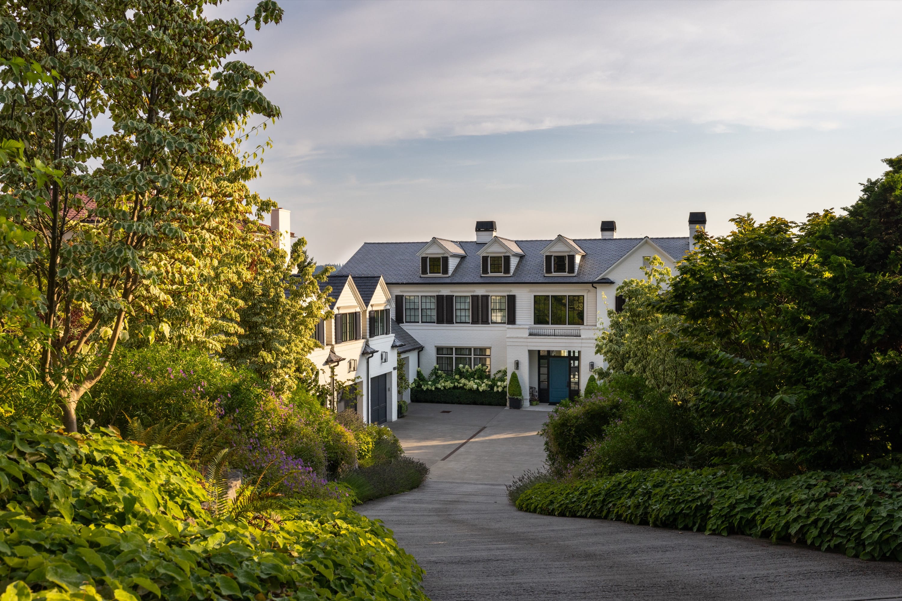 Large white home at end of tree-lined driveway with lush landscaping and soft evening light.
