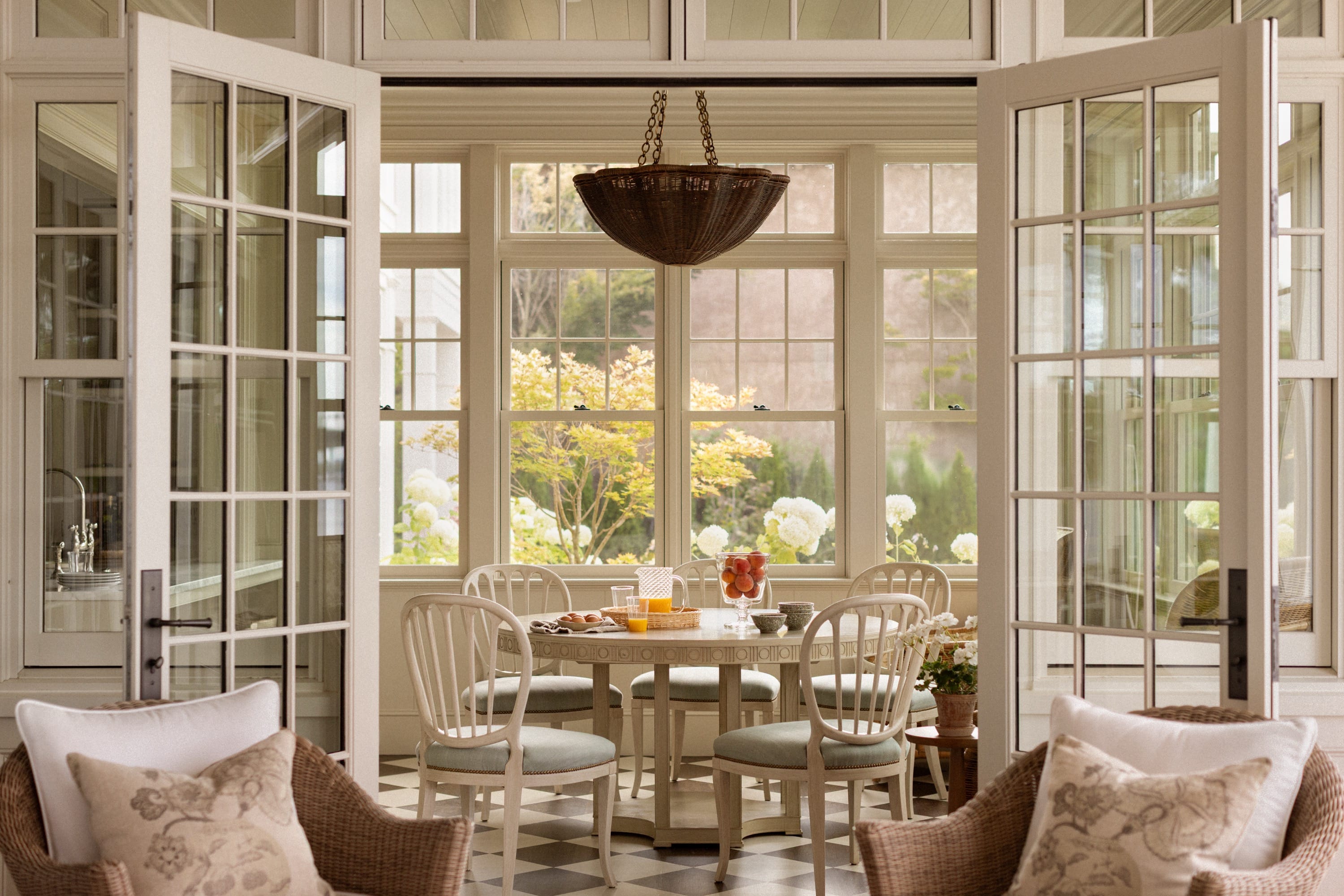 Sunlit dining area with round table, six white chairs, and glass doors opening to a garden with green and white flowers.